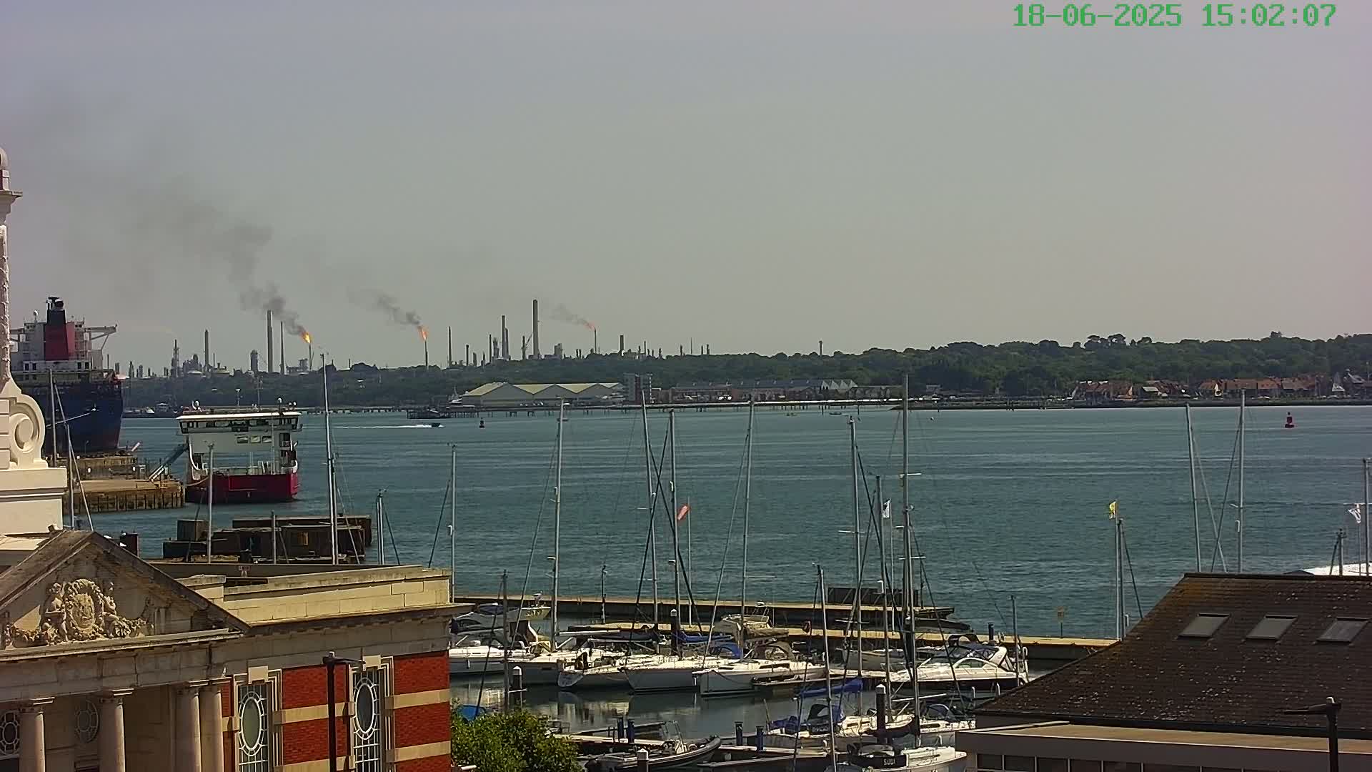 A harbor filled with sailboats and moored ships, with a distant industrial plant emitting smoke plumes under a hazy sky.