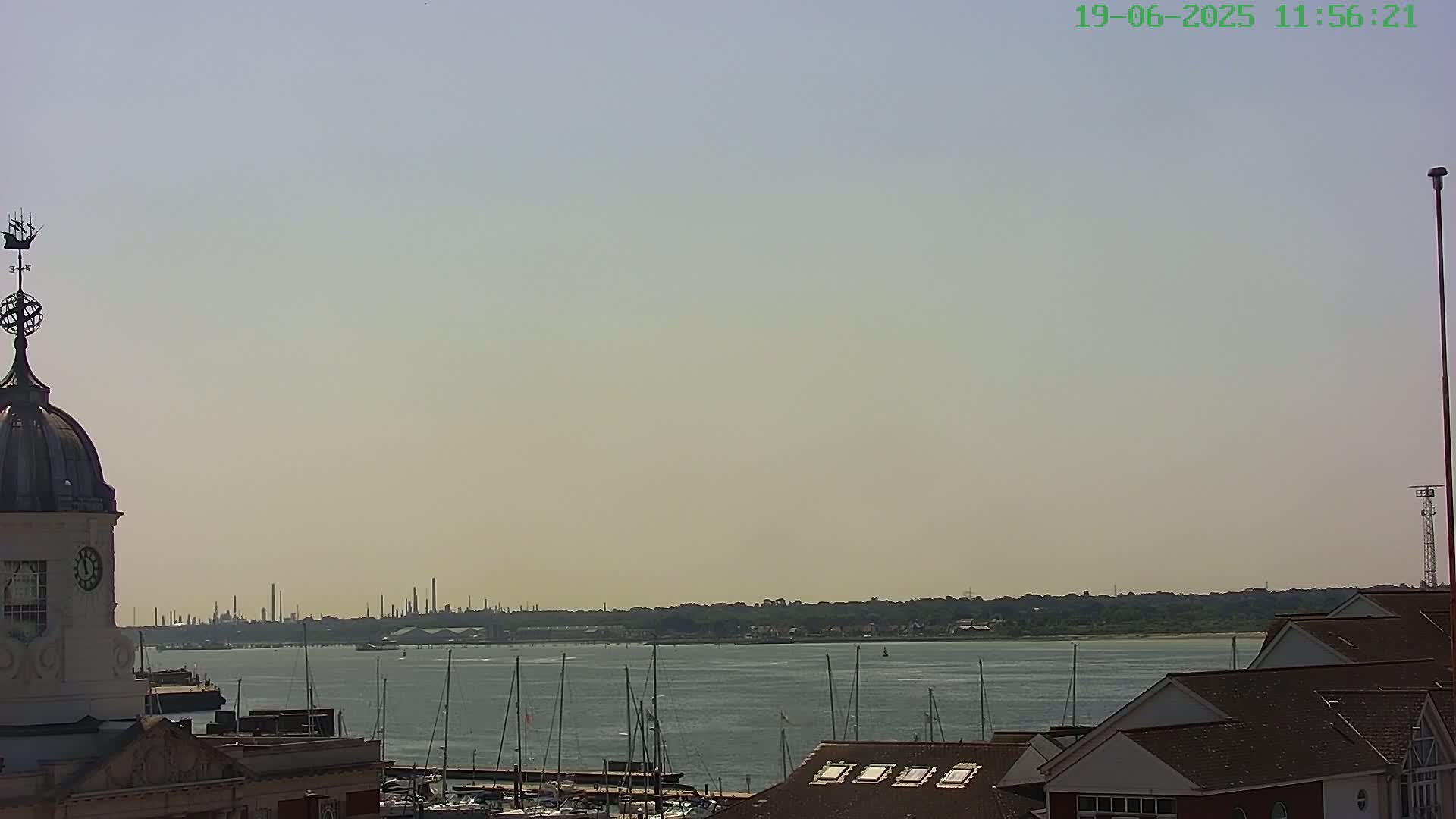 A hazy, sunny day overlooks a harbor filled with sailboats, with a distant industrial skyline and a building with a cupola in the foreground.