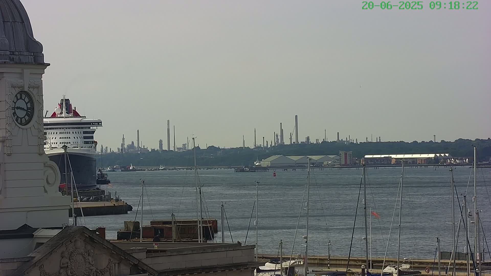 A large cruise ship is docked near a clock tower, with a hazy view of an industrial area and a marina across calm water.