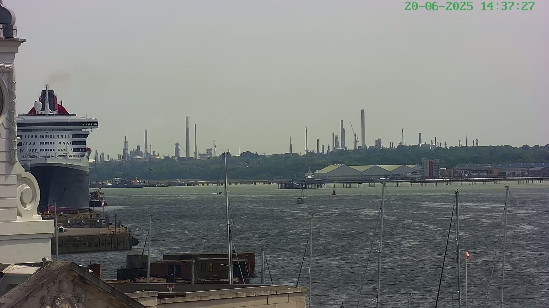 A large cruise ship is docked near a body of water, with an industrial plant visible in the distance on a hazy day.