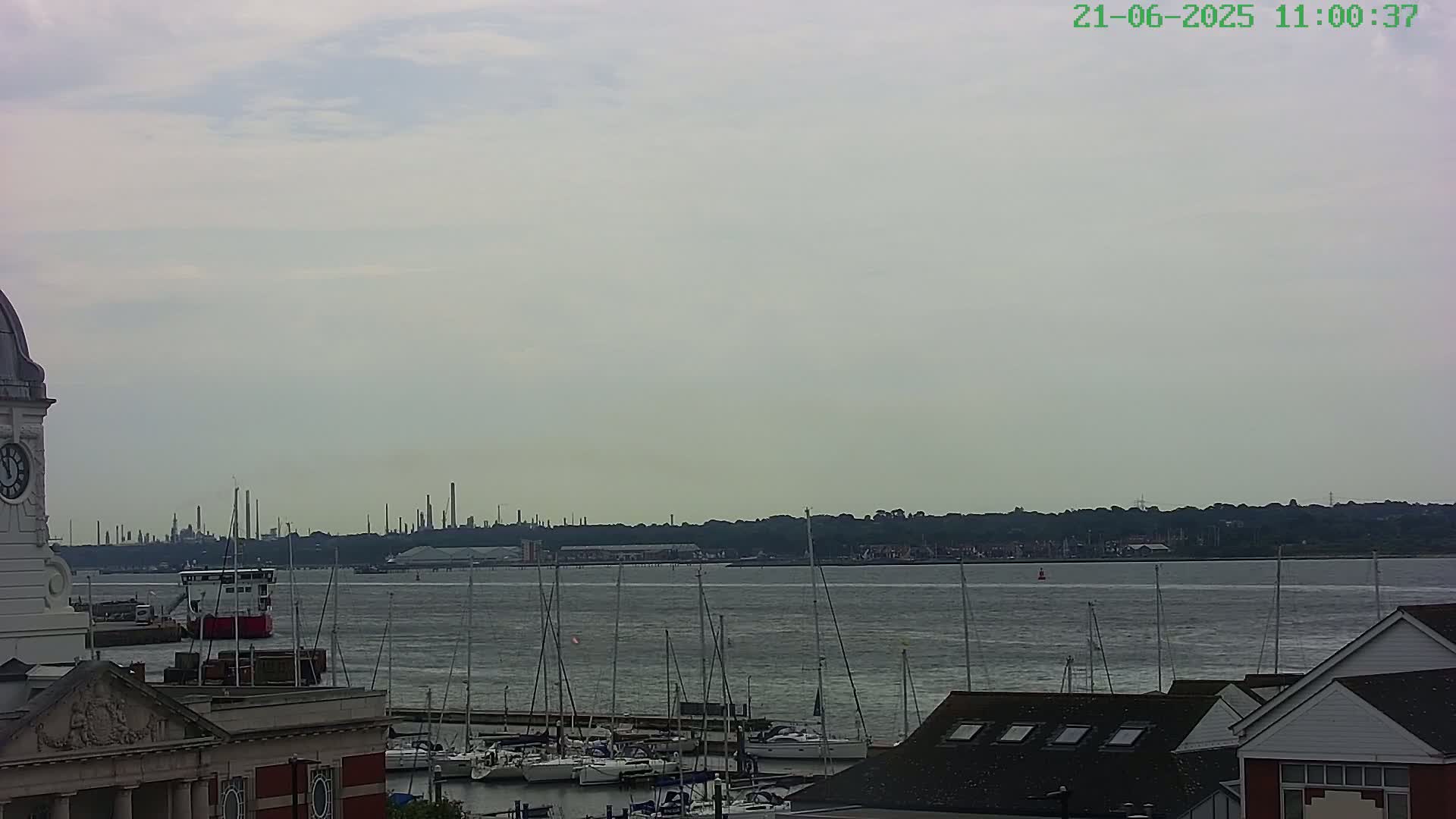 A waterfront scene on an overcast day shows a body of water with numerous sailboats docked, a distant industrial skyline, and a portion of a clock tower and building in the foreground.