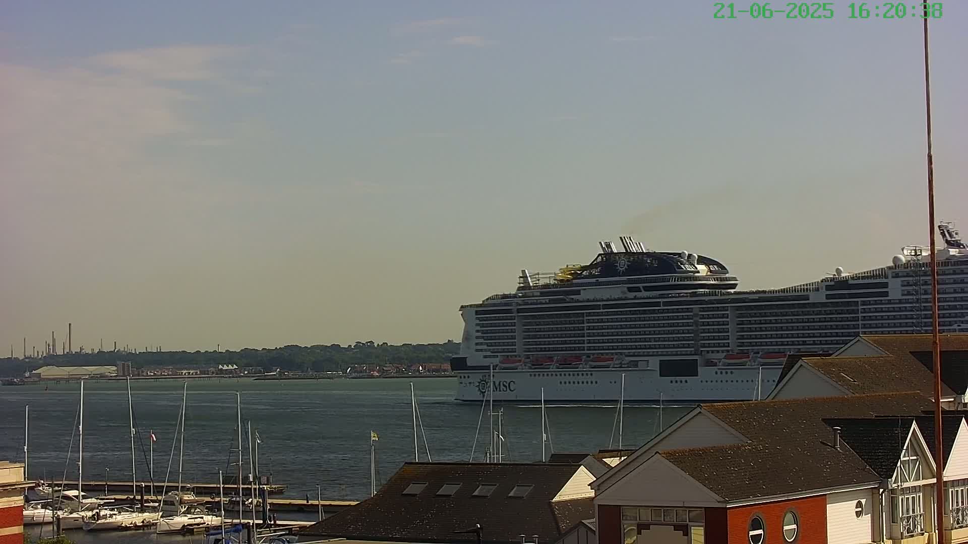 A large cruise ship is in the water under a partly cloudy sky, with smaller boats and buildings visible in the foreground.
