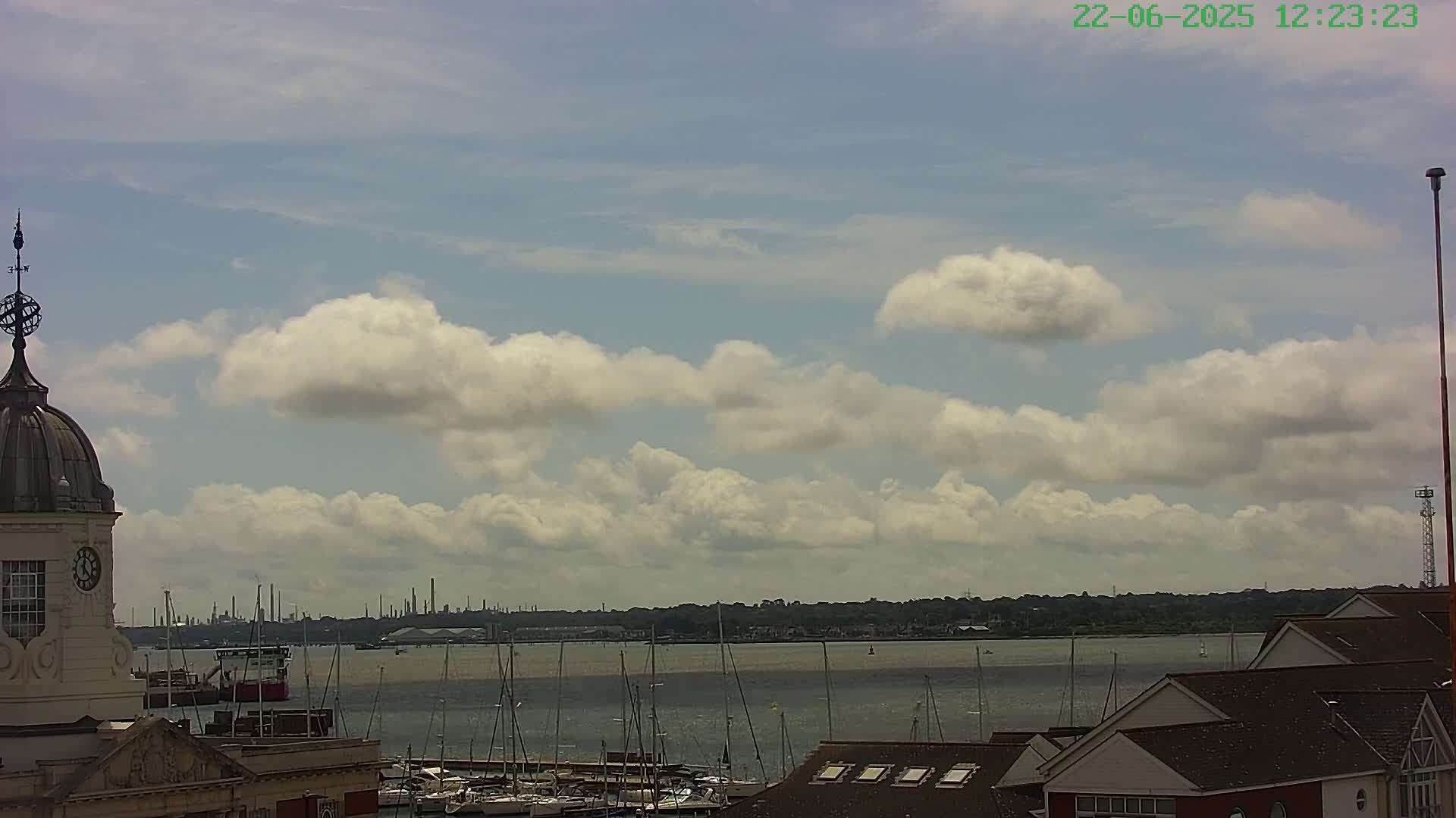 A partly cloudy sky overlooks a harbor filled with sailboats, with a distant industrial skyline visible across the water and a clock tower in the foreground.