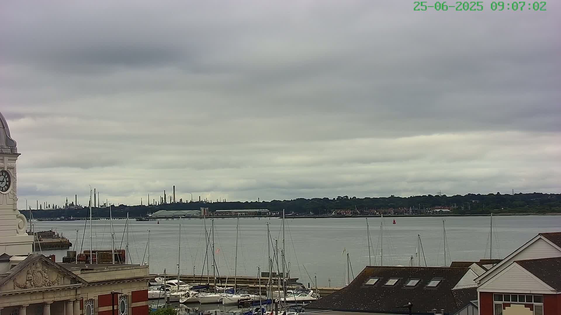 A waterfront scene under a cloudy sky shows a marina with numerous sailboats, a distant industrial skyline, and a portion of a building with a clock tower in the foreground.