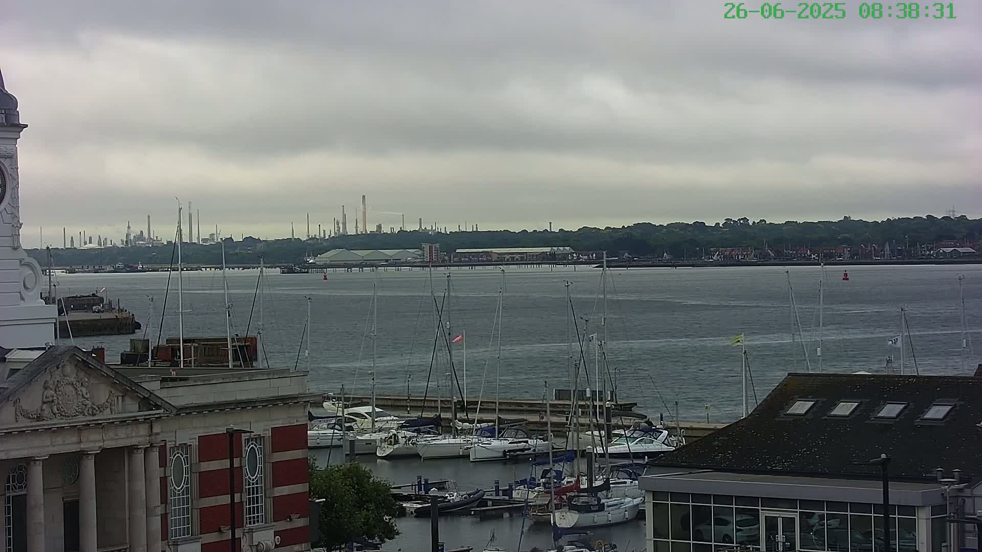 A harbor filled with sailboats is situated next to a brick building with a clock tower, under an overcast sky with a distant industrial plant visible across the water.