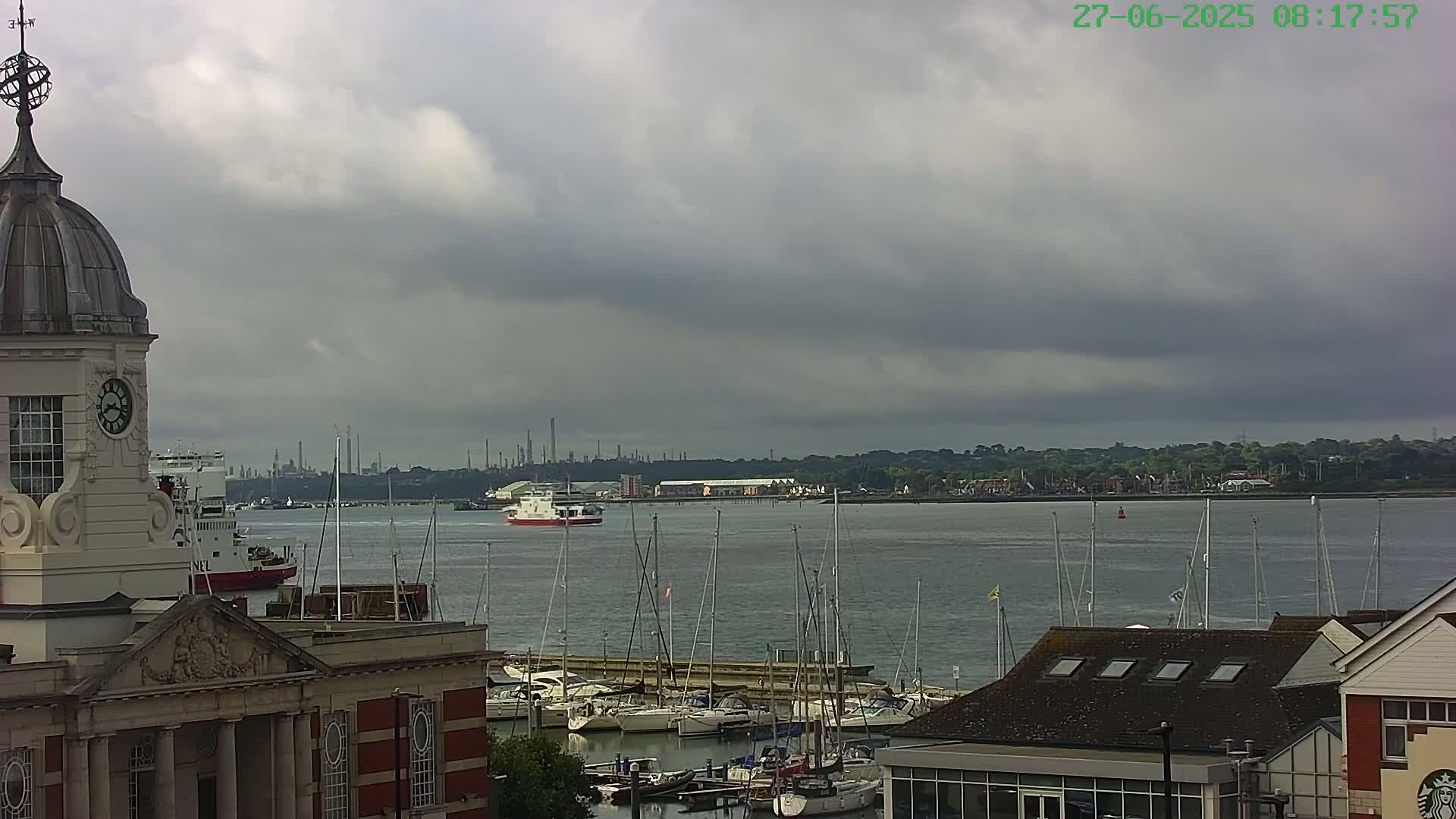 A harbor with numerous sailboats is seen from atop a clock tower building under a cloudy sky, with a large ferry in the distance and an industrial area visible on the horizon.