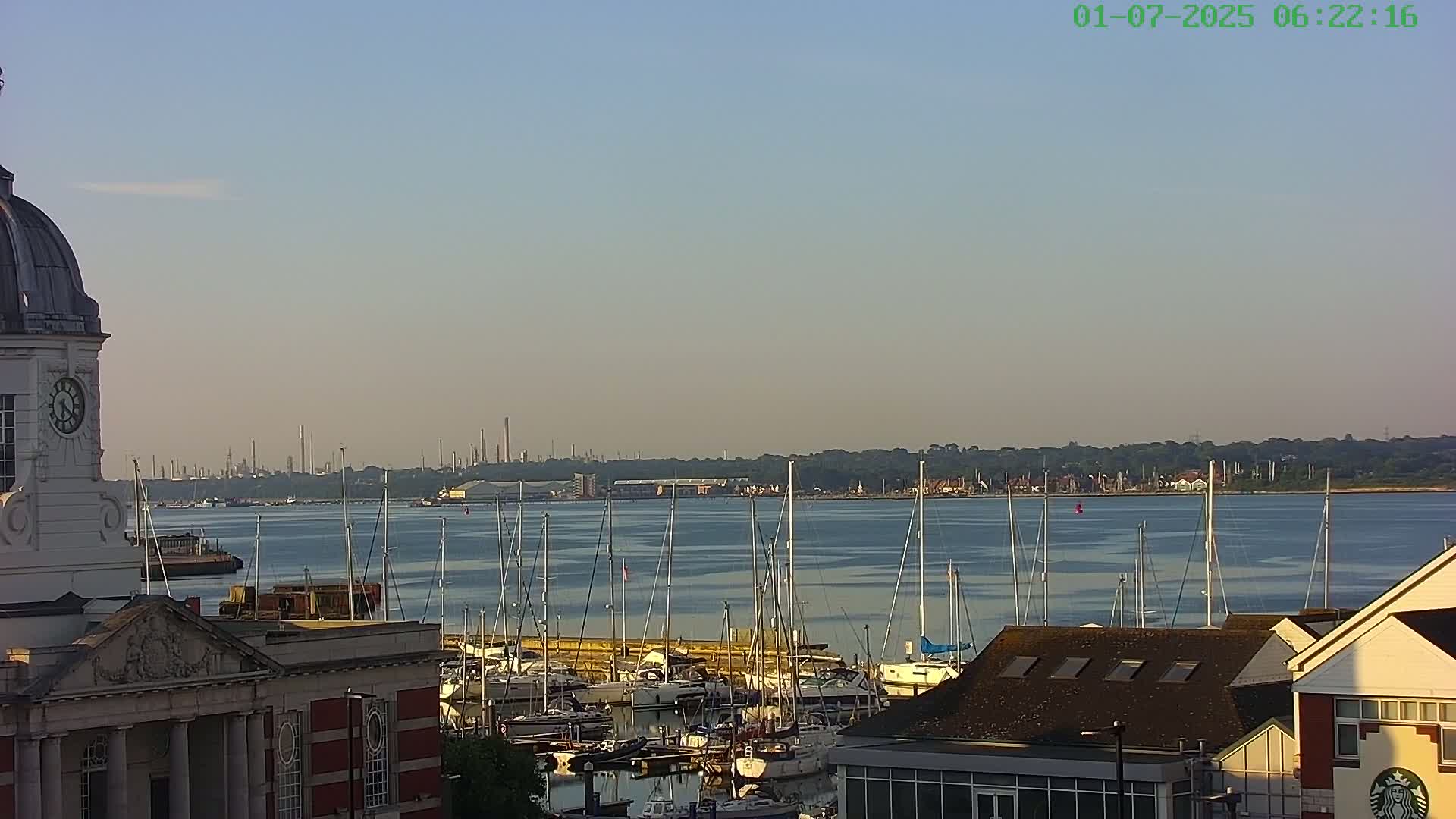 A calm, clear morning reveals a marina filled with sailboats, a waterfront building with a clock tower, and a distant industrial skyline.