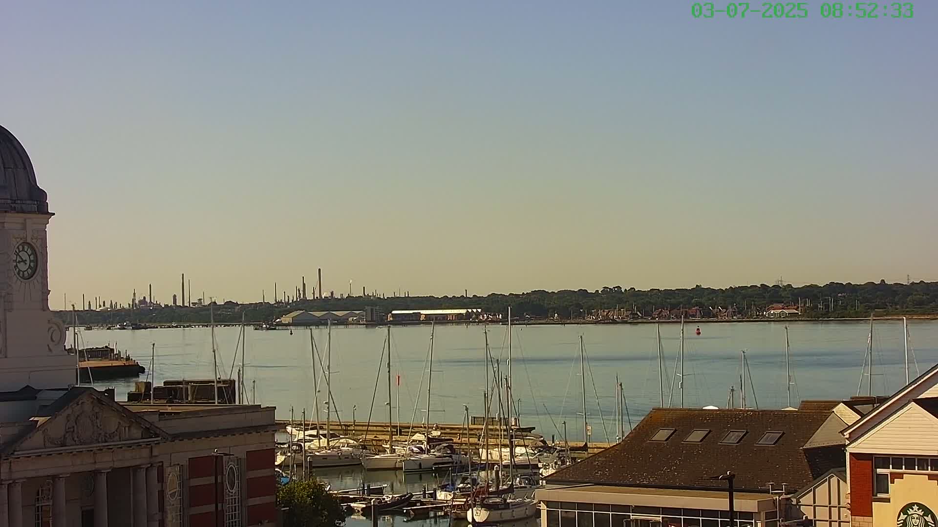 A harbor filled with sailboats is seen from an elevated vantage point, with a clock tower to the left and industrial structures visible in the distance under a clear sky.