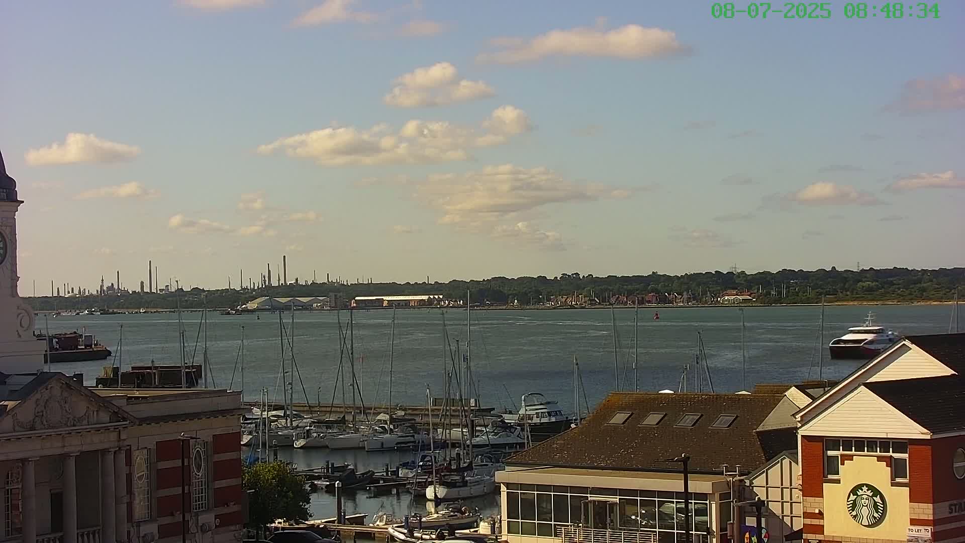 A harbor filled with sailboats and yachts sits under a partly cloudy sky, with an industrial area visible in the distance across the water.