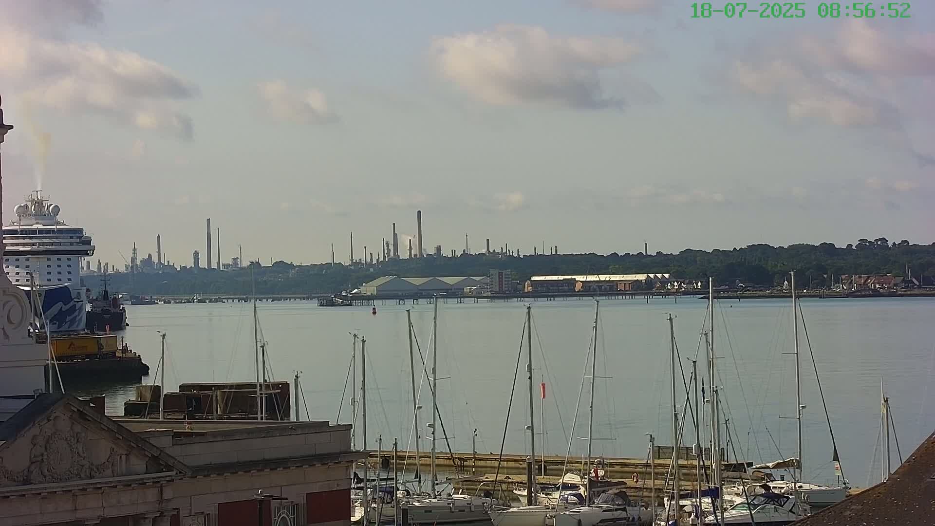A cruise ship is docked near a marina full of sailboats, with an industrial plant visible in the distance across a calm body of water under a partly cloudy sky.