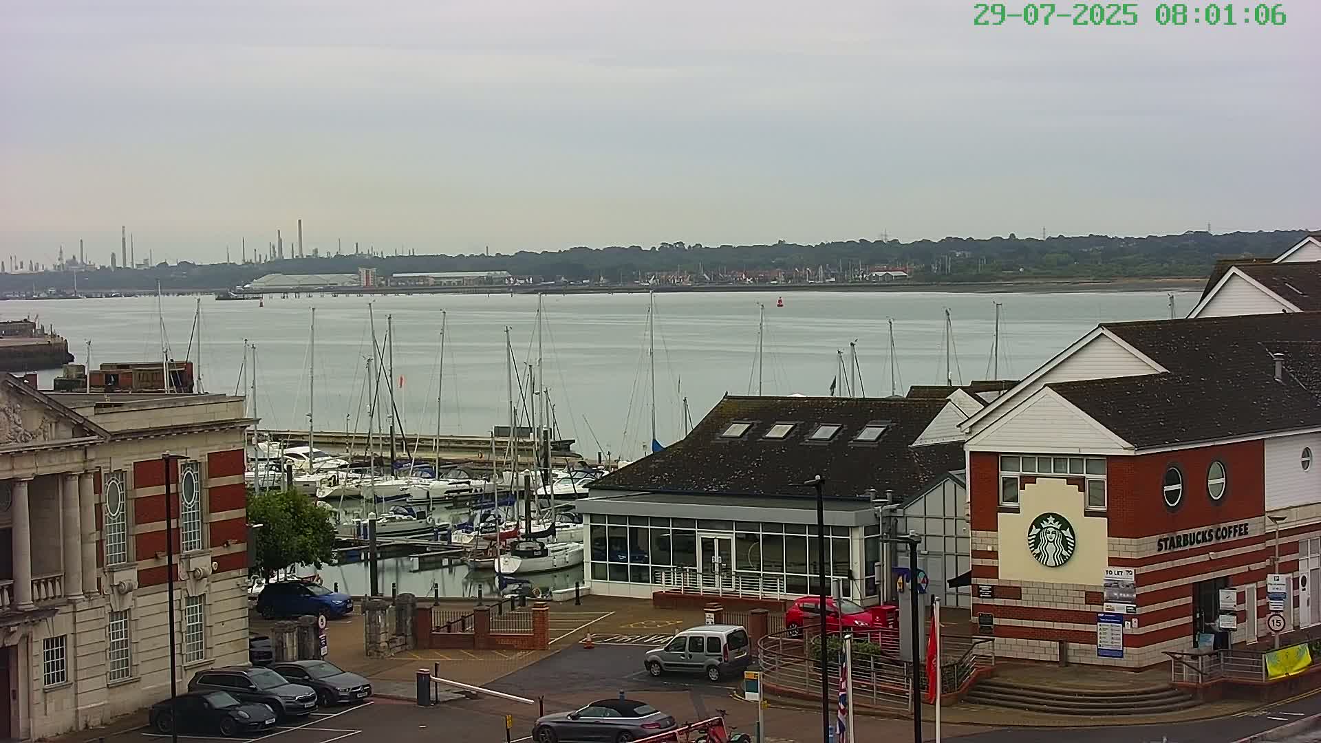 A marina filled with sailboats is visible near a brick building with a Starbucks, under an overcast sky, with an industrial area in the background across a body of water.