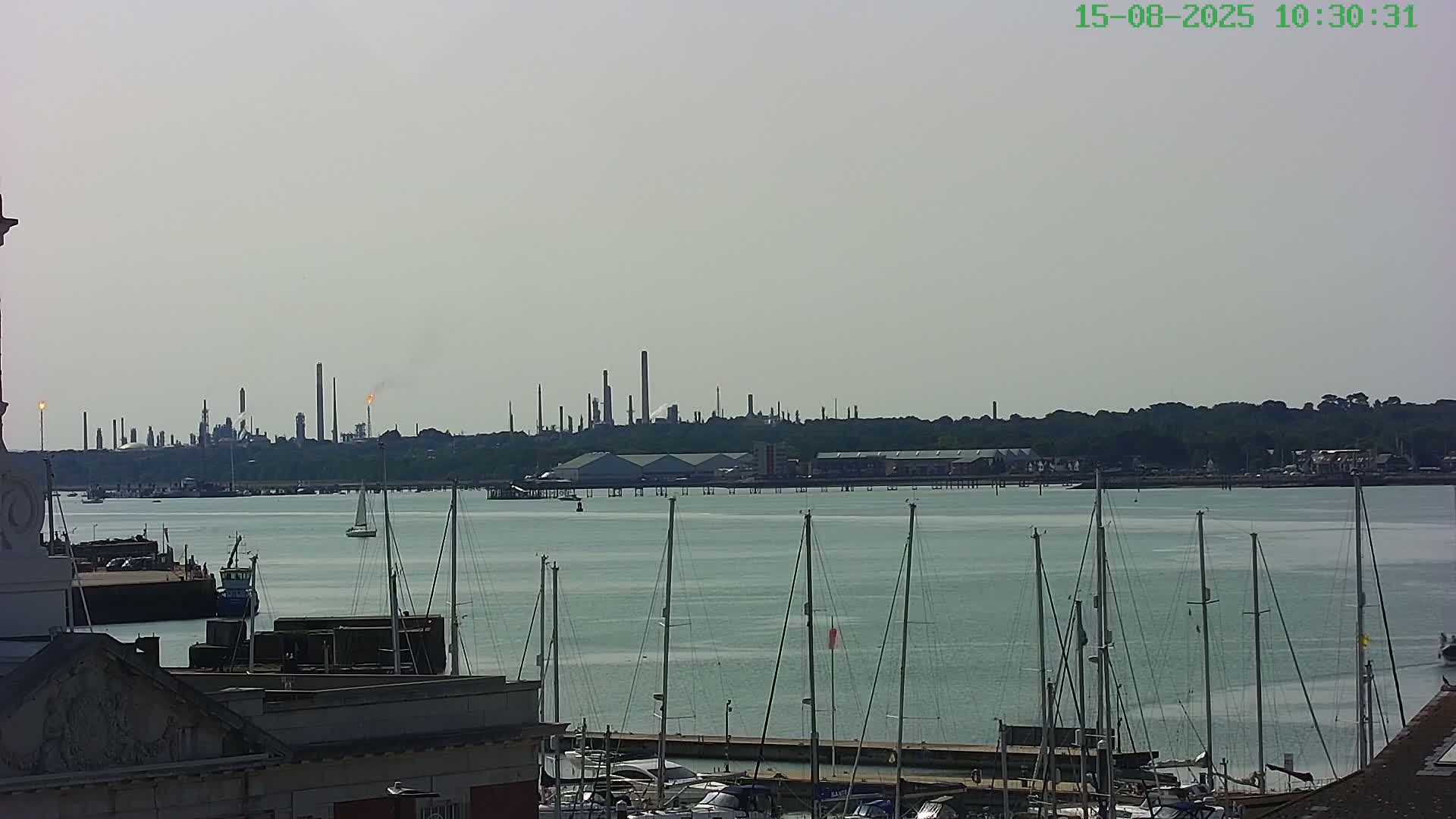 A marina filled with sailboats is visible near a brick building with a Starbucks, under an overcast sky, with an industrial area in the background across a body of water.