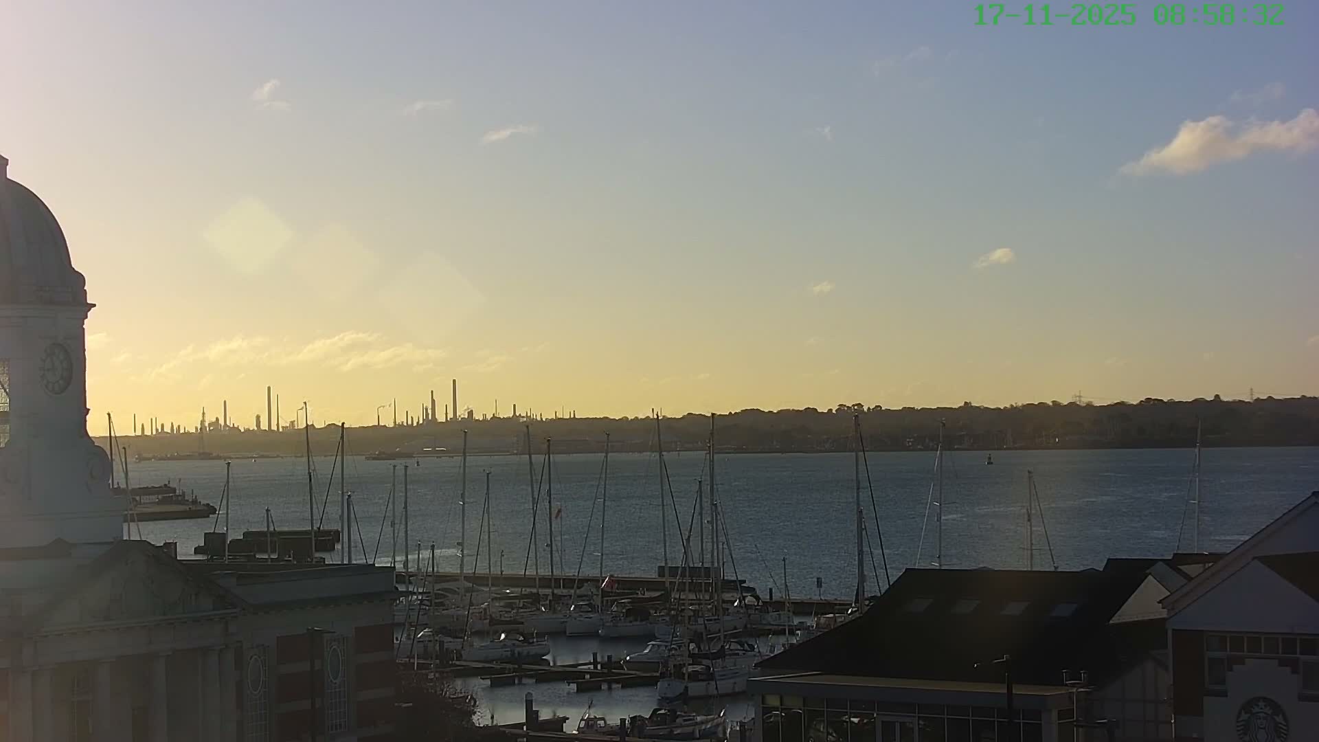 On a clear morning with a golden sunrise hue, a bustling harbor features a domed clock tower in the foreground, numerous sailboats docked in the calm waters, and a distant industrial skyline across the bay.