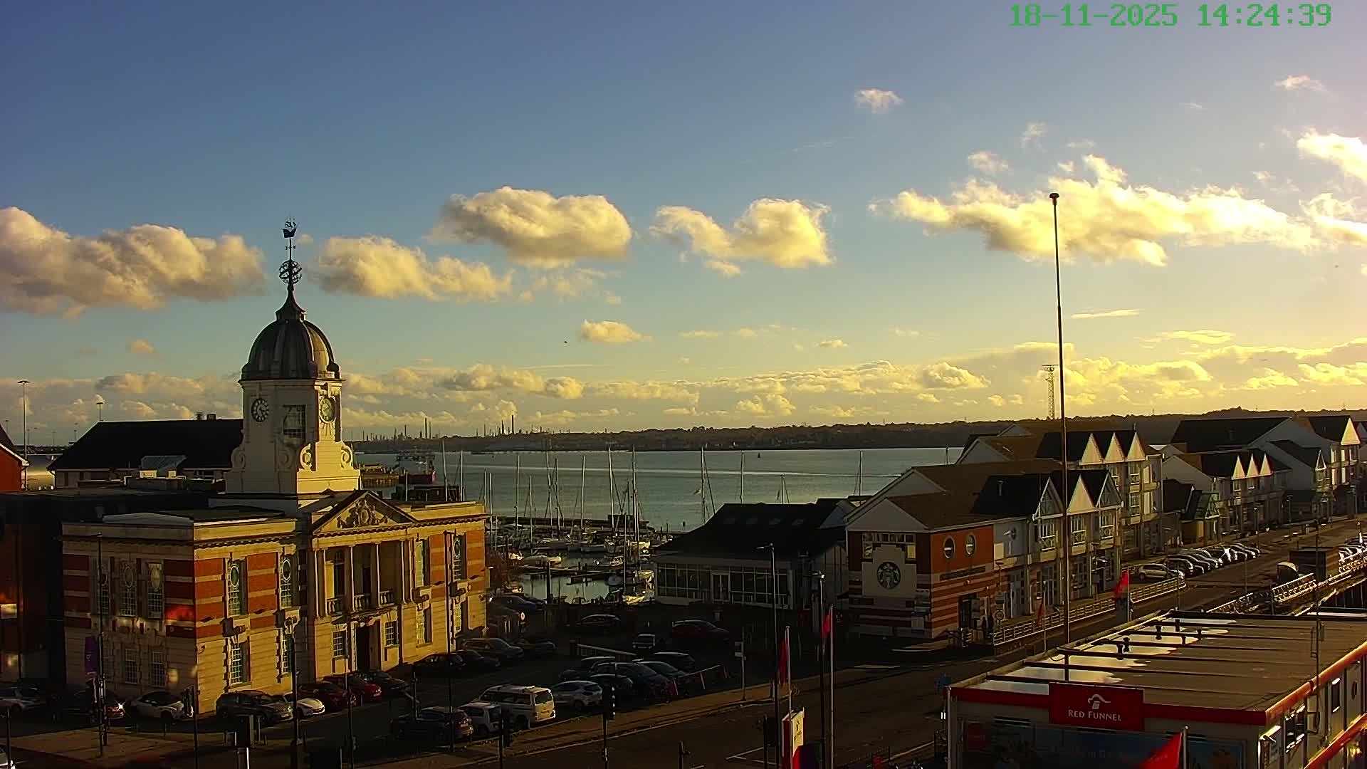 On a clear morning with a golden sunrise hue, a bustling harbor features a domed clock tower in the foreground, numerous sailboats docked in the calm waters, and a distant industrial skyline across the bay.