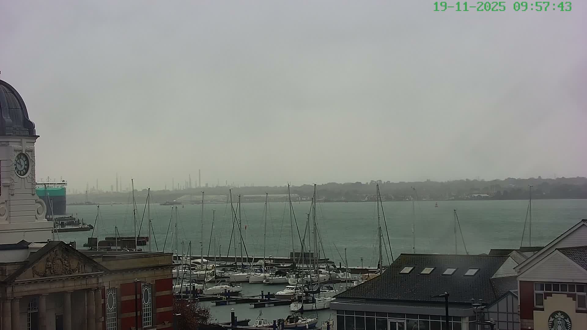 A hazy and overcast day casts a muted light over a bustling marina filled with numerous sailboats, with a large green ship navigating the choppy water, a prominent clock tower building on the left, and industrial structures visible on the distant shore.