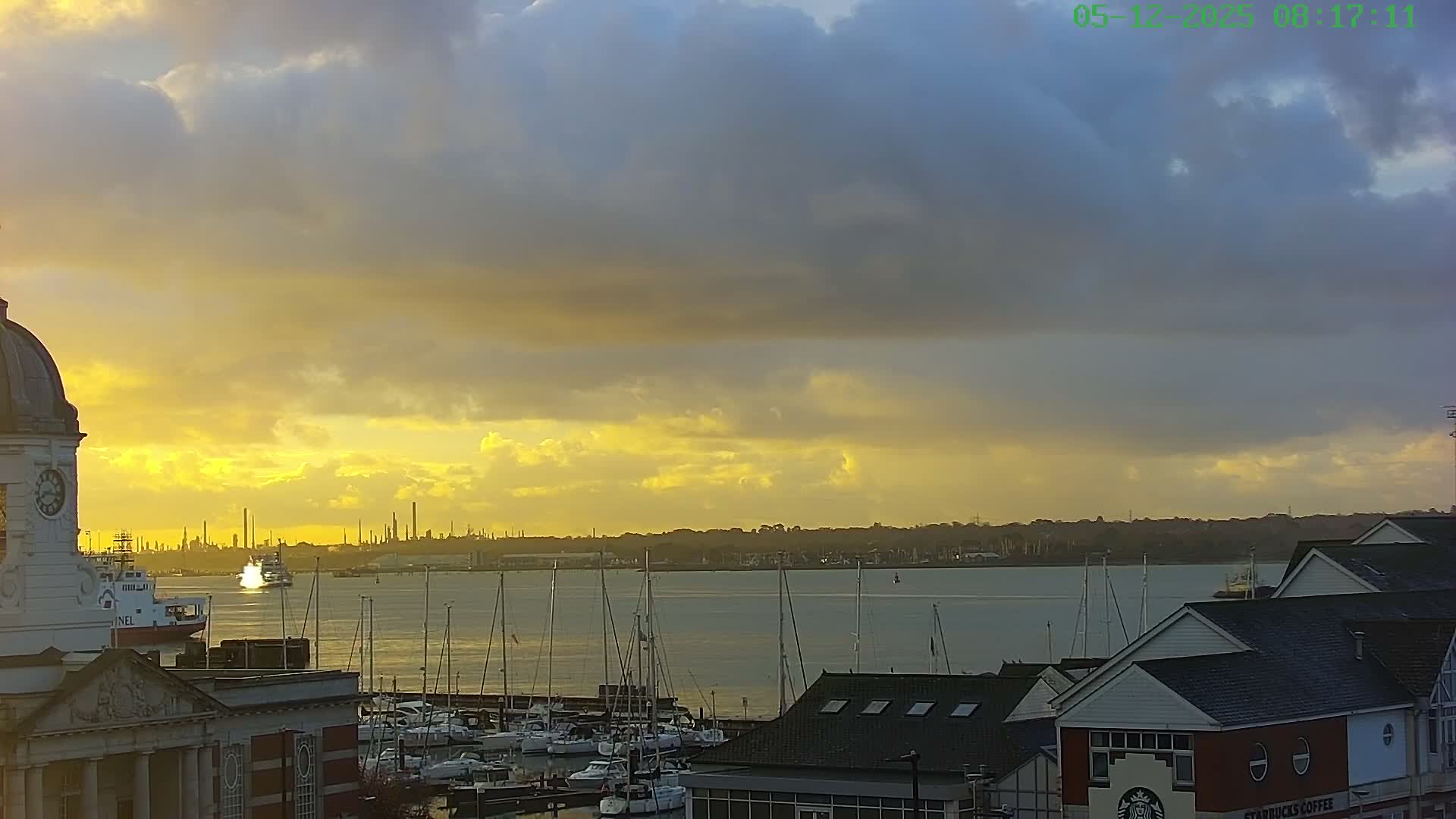 A dramatic sunrise casts a golden glow across a bustling harbor, illuminating numerous sailboats, a prominent clock tower, and distant industrial silhouettes under a sky filled with dark, heavy clouds interspersed with bright yellow hues.