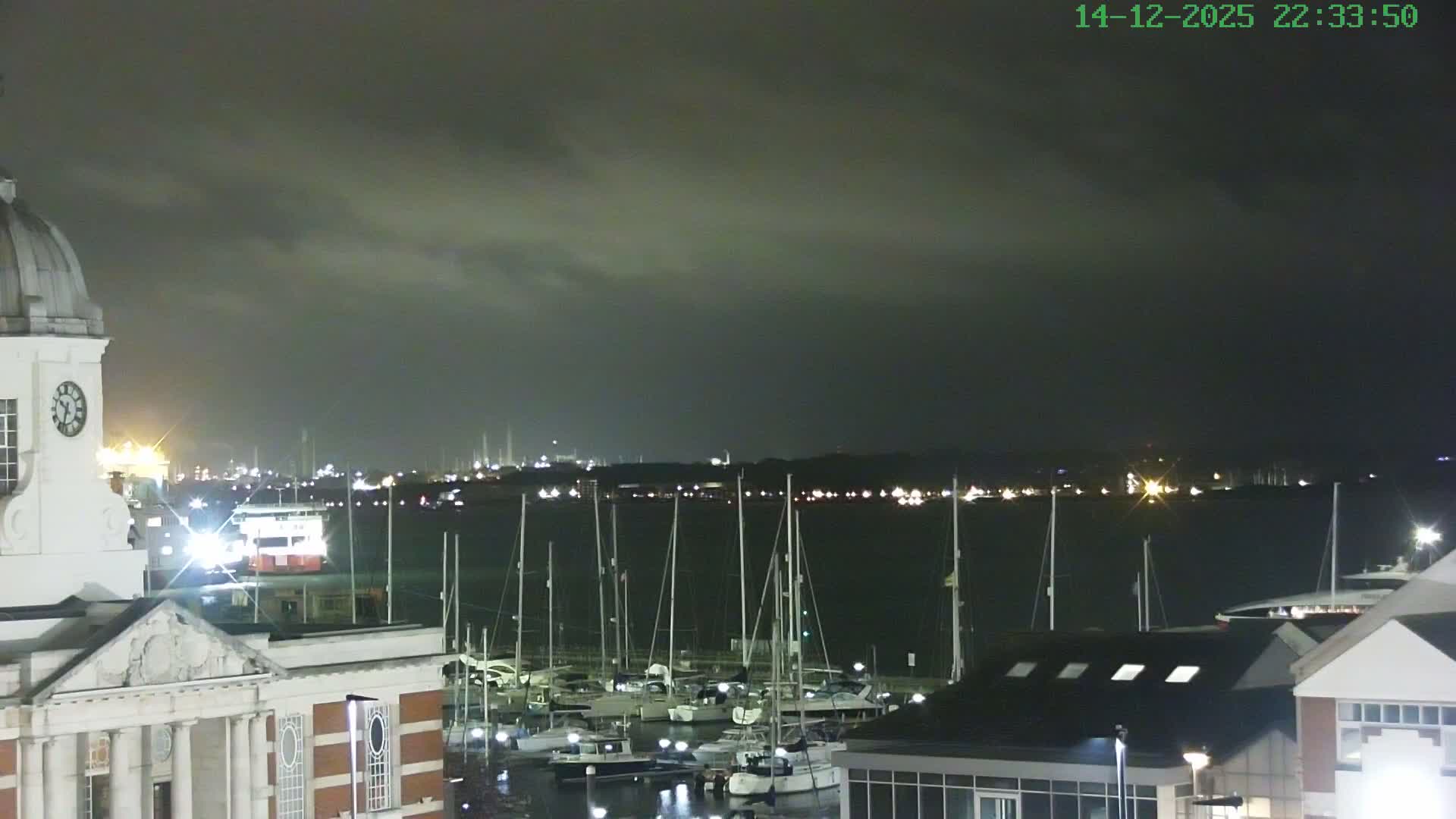 A dramatic sunrise casts a golden glow across a bustling harbor, illuminating numerous sailboats, a prominent clock tower, and distant industrial silhouettes under a sky filled with dark, heavy clouds interspersed with bright yellow hues.