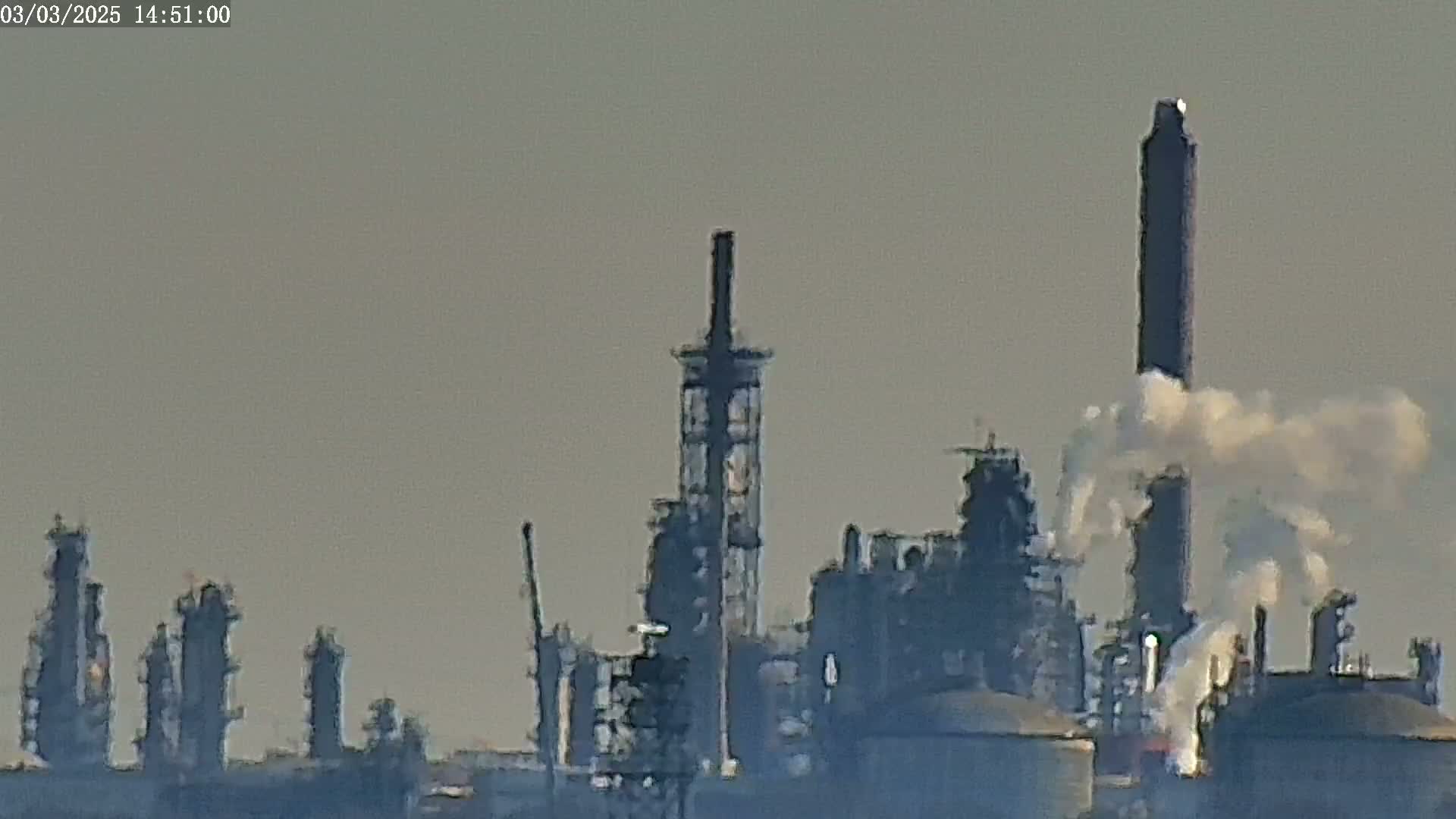 A hazy industrial complex with numerous tall structures and smoke plumes emitting from several stacks is visible under a muted sky.