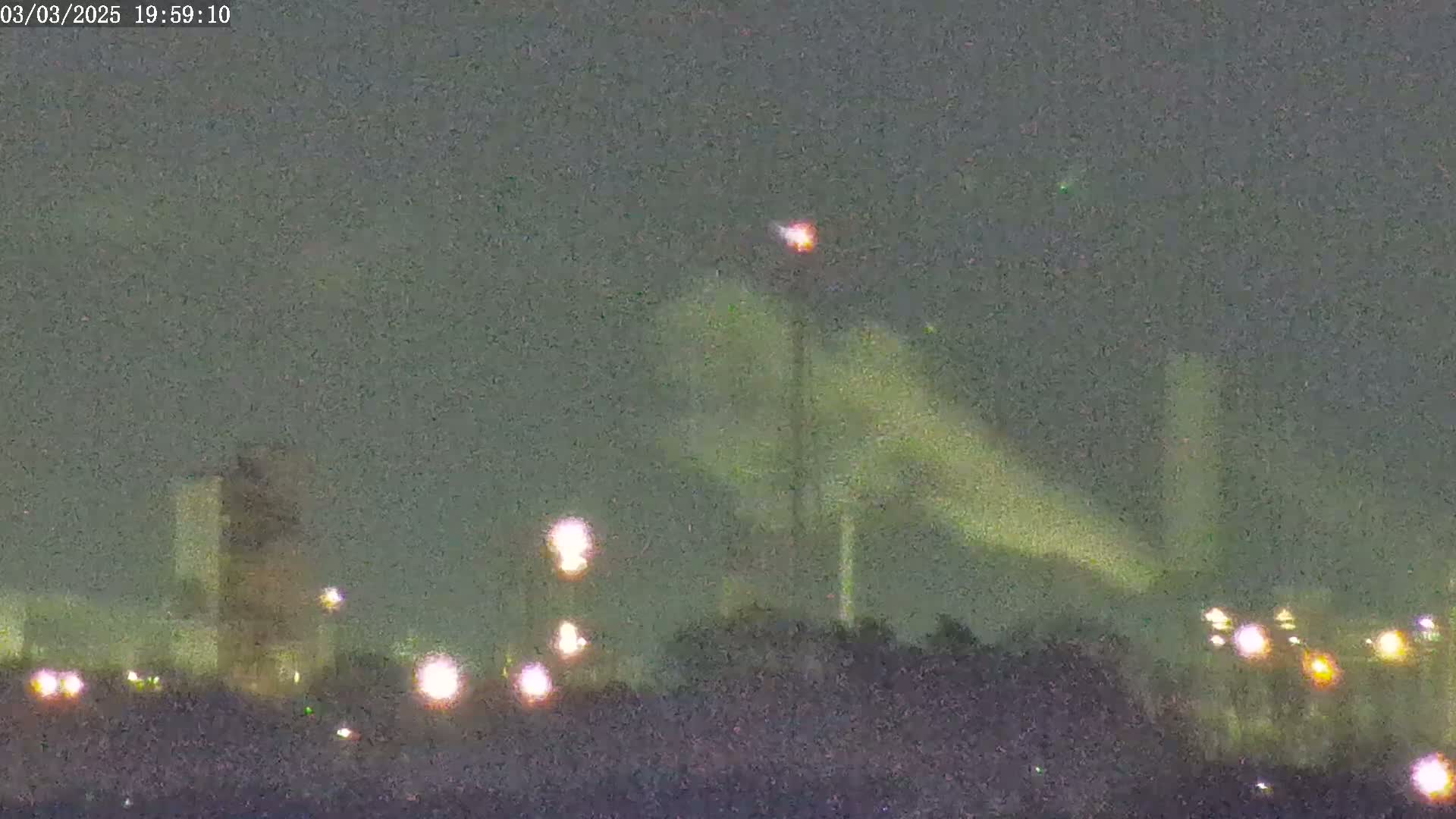 A nighttime view of an industrial area with several tall structures emitting plumes of light-greenish smoke under a dark, overcast sky.