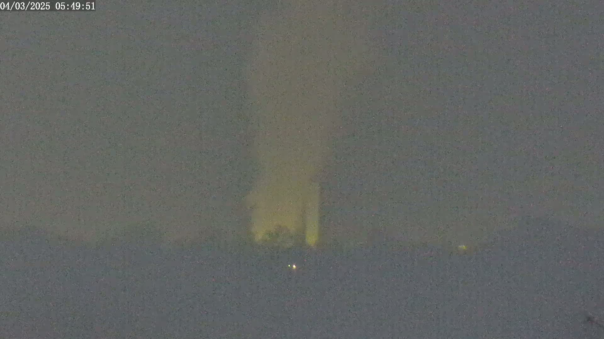 A dimly lit, hazy scene at night shows a faint, yellowish glow emanating from a structure in the distance.