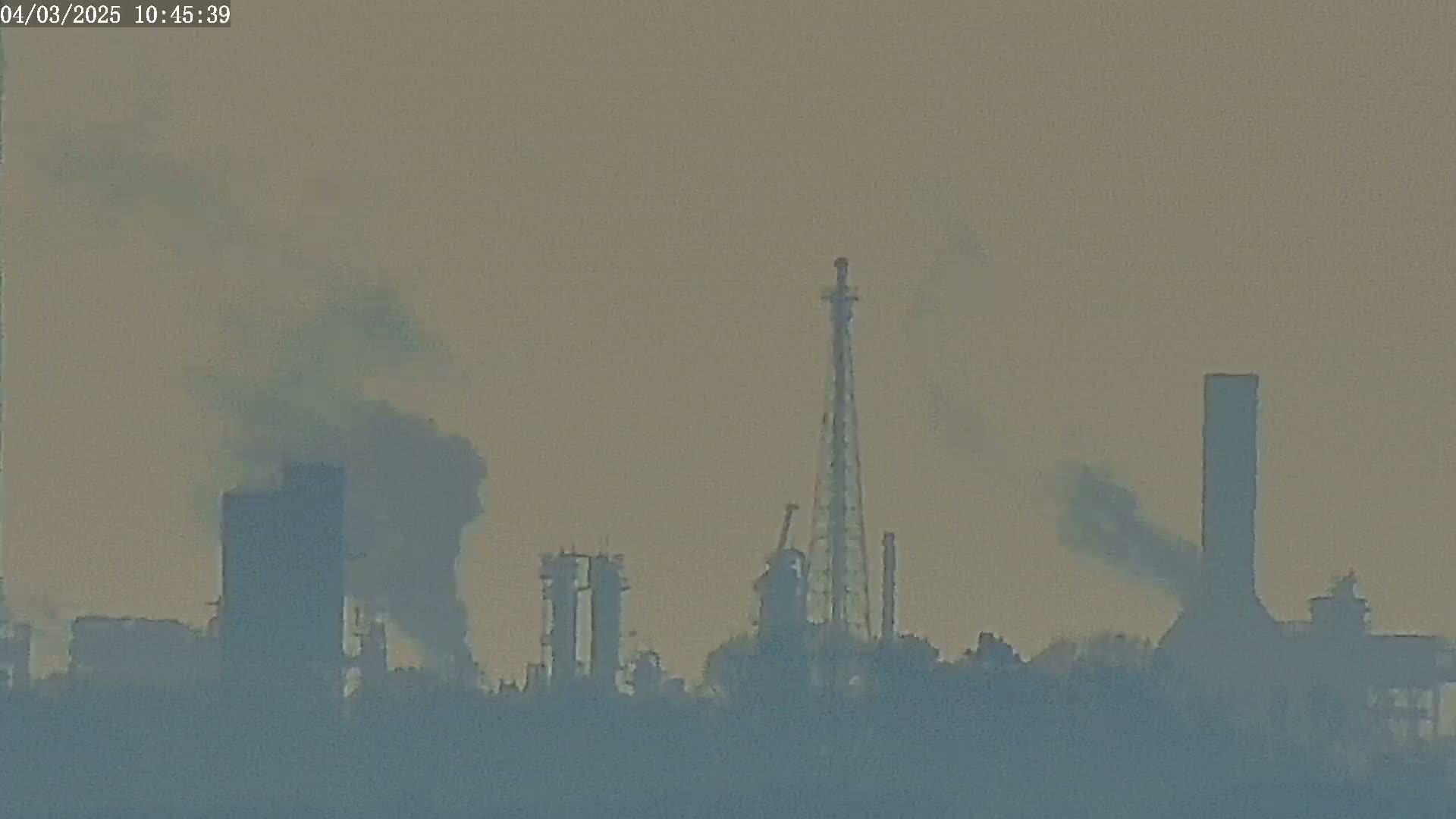 A hazy industrial complex with several tall structures and smoke plumes visible against a muted sky.