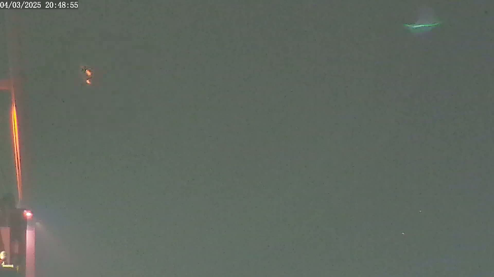 A nighttime, muted green-grey sky shows a bright orange vertical light and two small, faint lights, one of which is elongated and pale green.