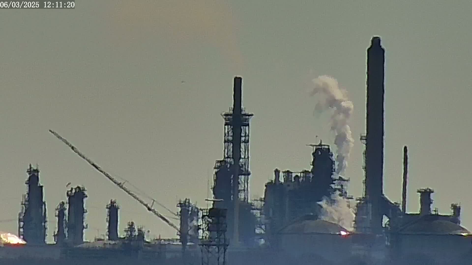 A large industrial complex with numerous tall structures and smokestacks emits white smoke under a hazy sky.