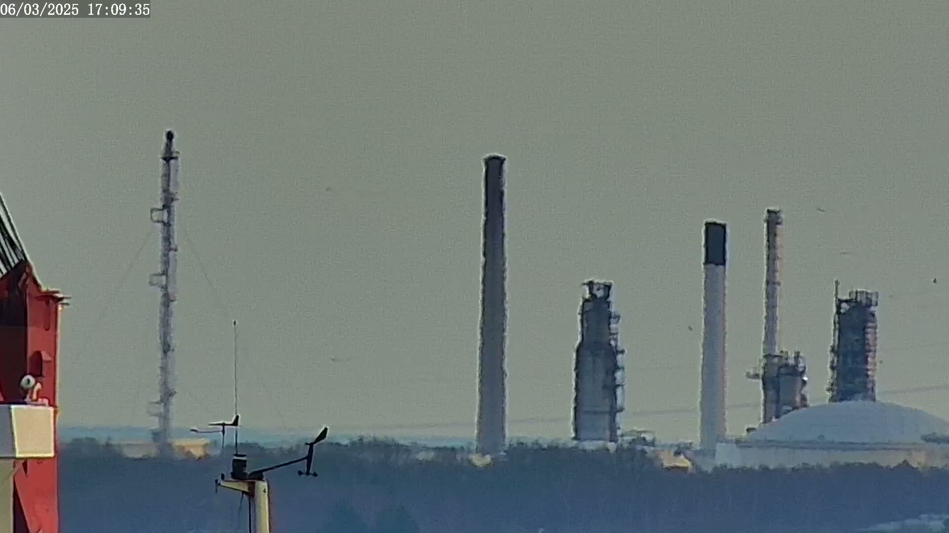 A hazy view shows several industrial smokestacks and refinery structures behind a weather station on a ship or dock.