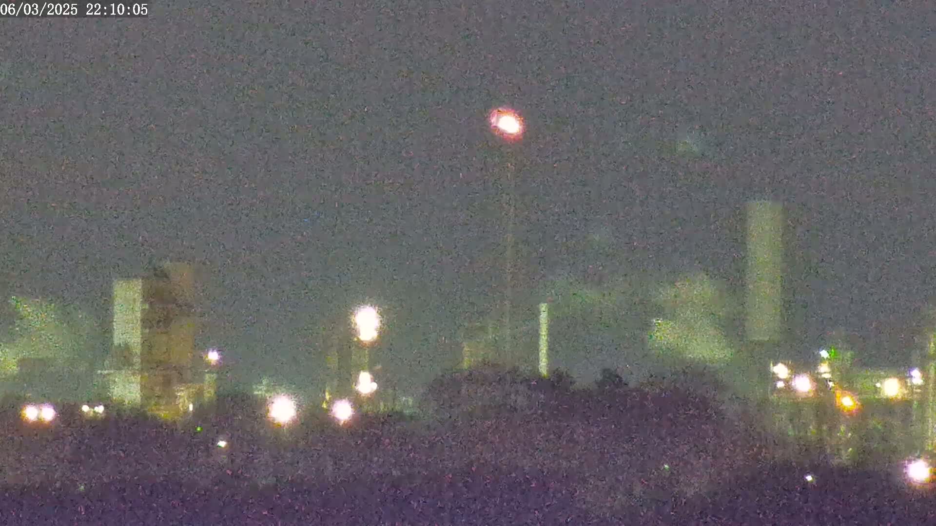 A nighttime, blurry view shows an industrial complex with several tall structures and numerous lights, under a dark, overcast sky.