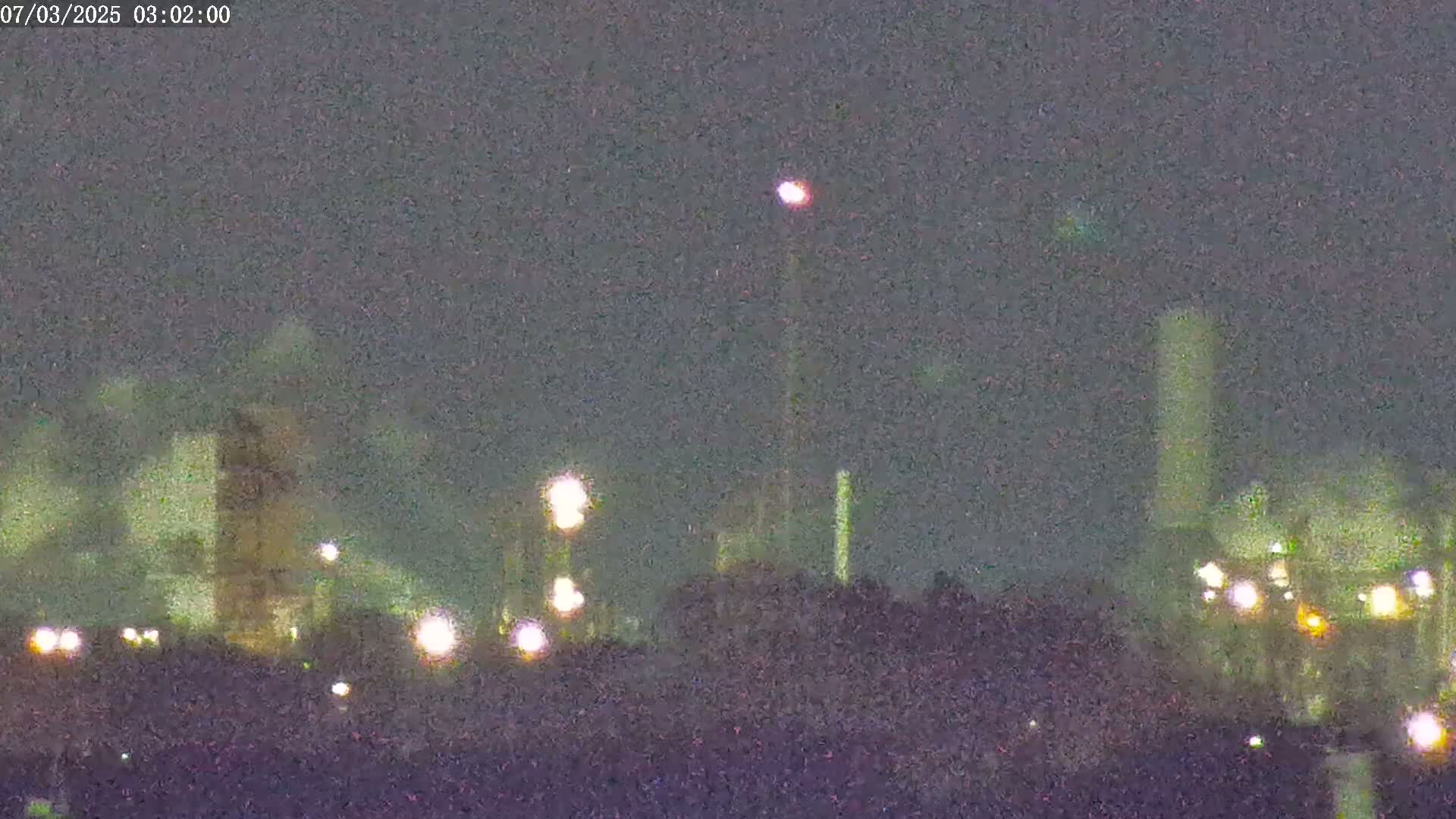 A nighttime long shot shows an industrial plant with illuminated structures and tall smokestacks under a dark, overcast sky.