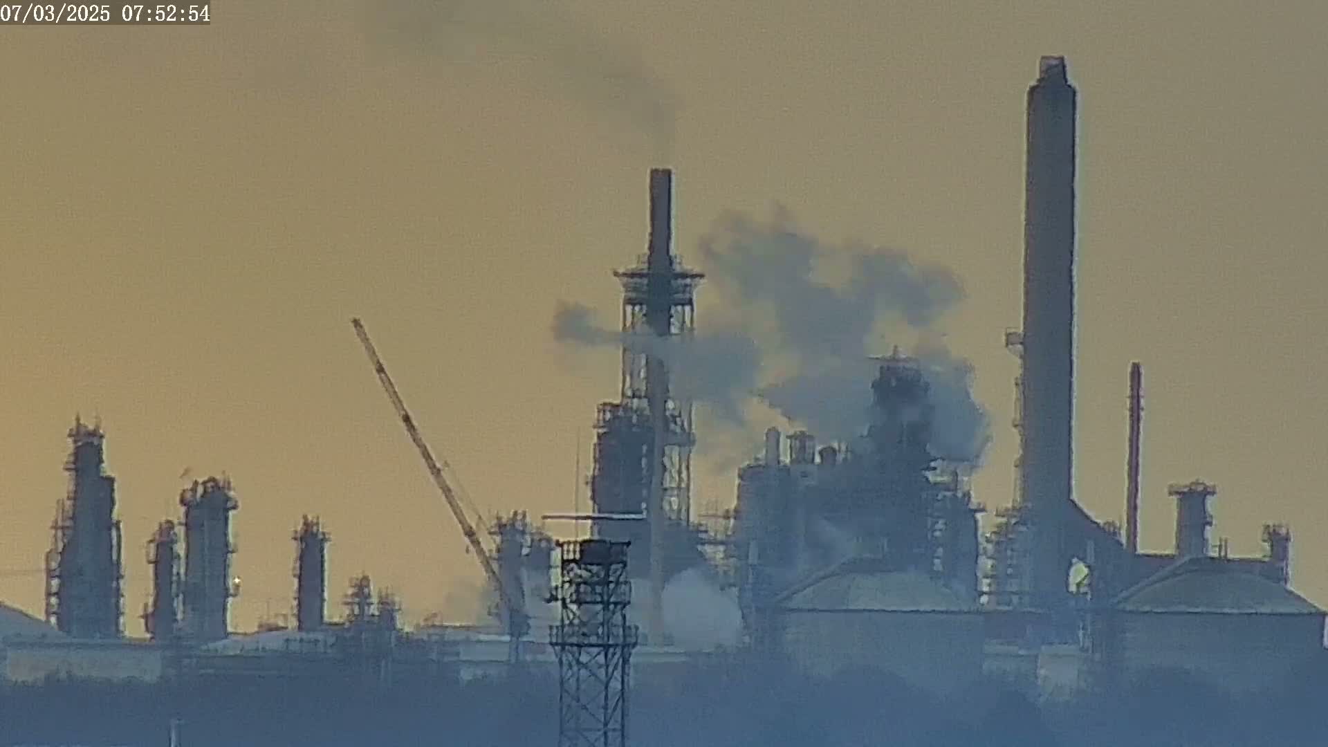 A hazy, pale-orange sunrise illuminates a large industrial complex emitting smoke from numerous tall structures, including several smokestacks and refinery towers, with a crane visible in the distance.