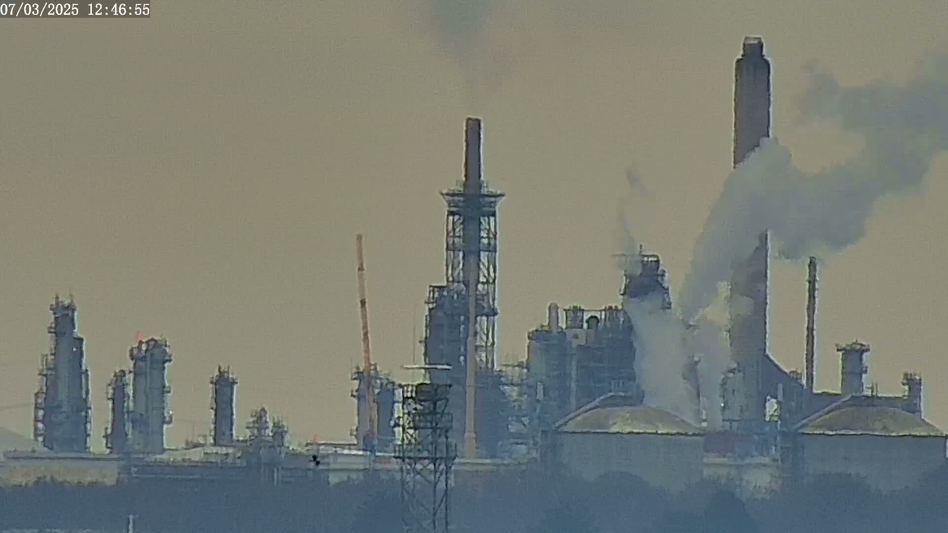 A hazy industrial complex with several tall smokestacks emitting smoke into a muted sky.