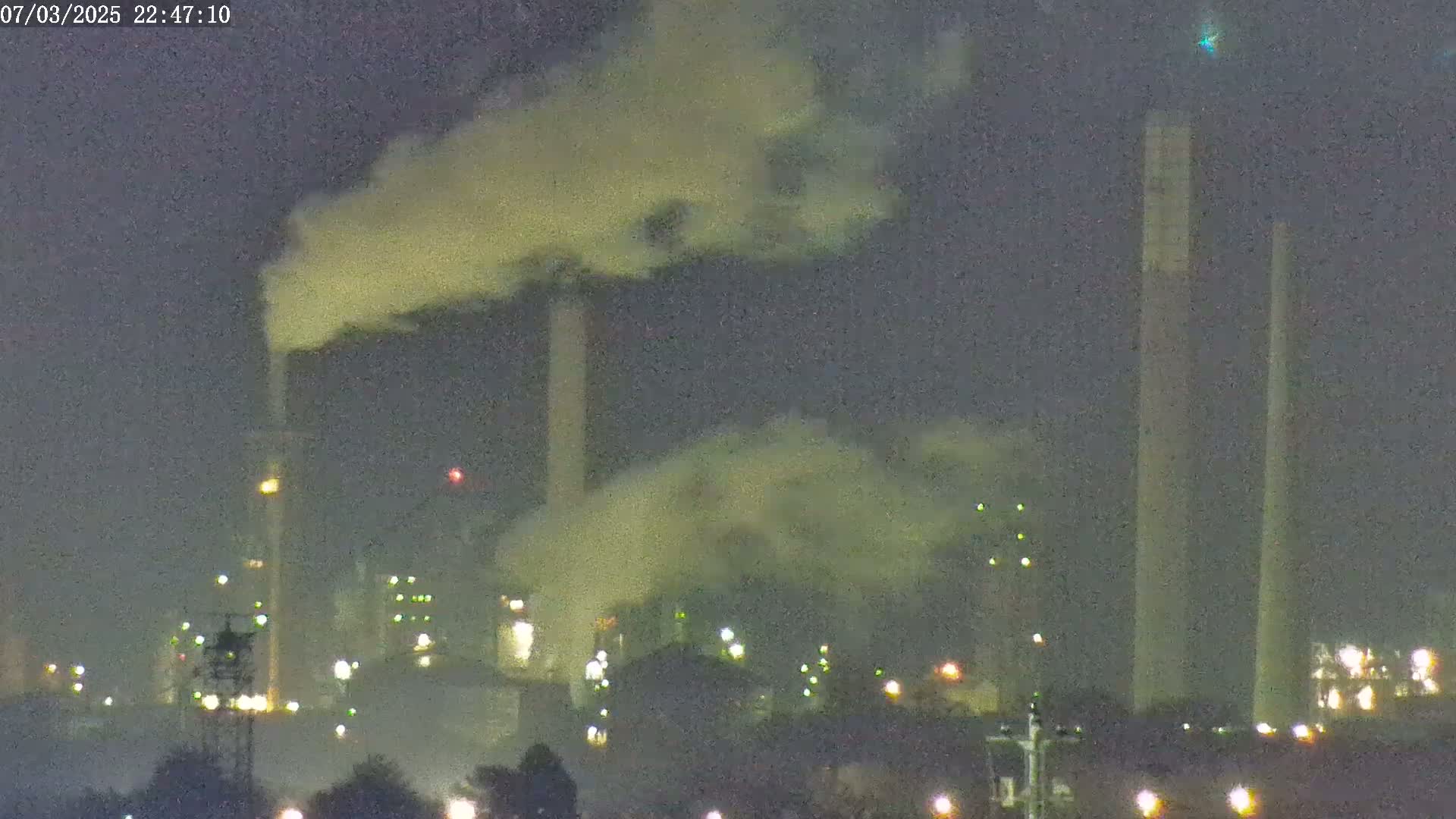 At night, an industrial plant with tall smokestacks emitting plumes of smoke is illuminated by various lights.