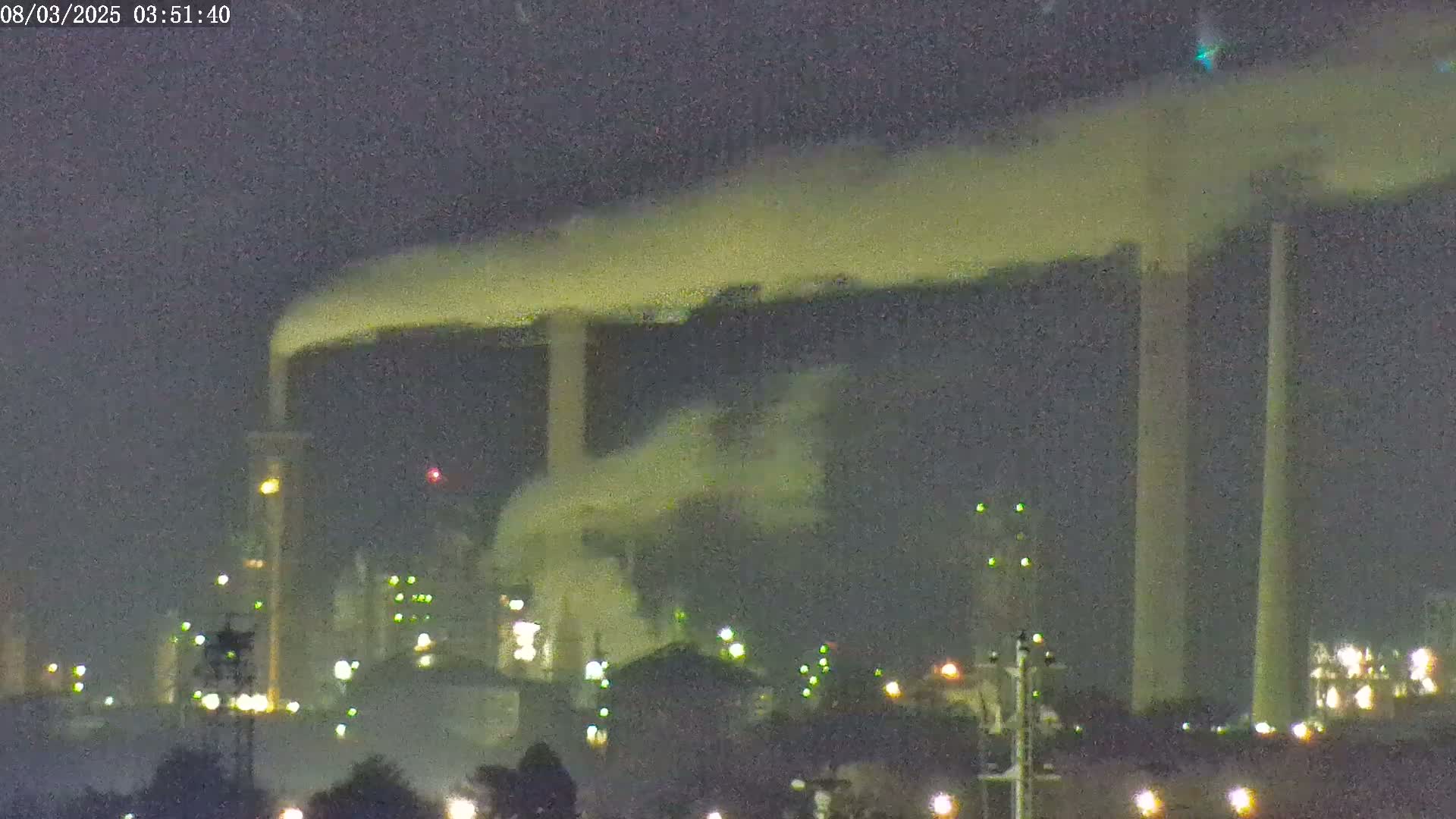 At night, a large industrial plant emits a plume of smoke or steam into a dark sky.