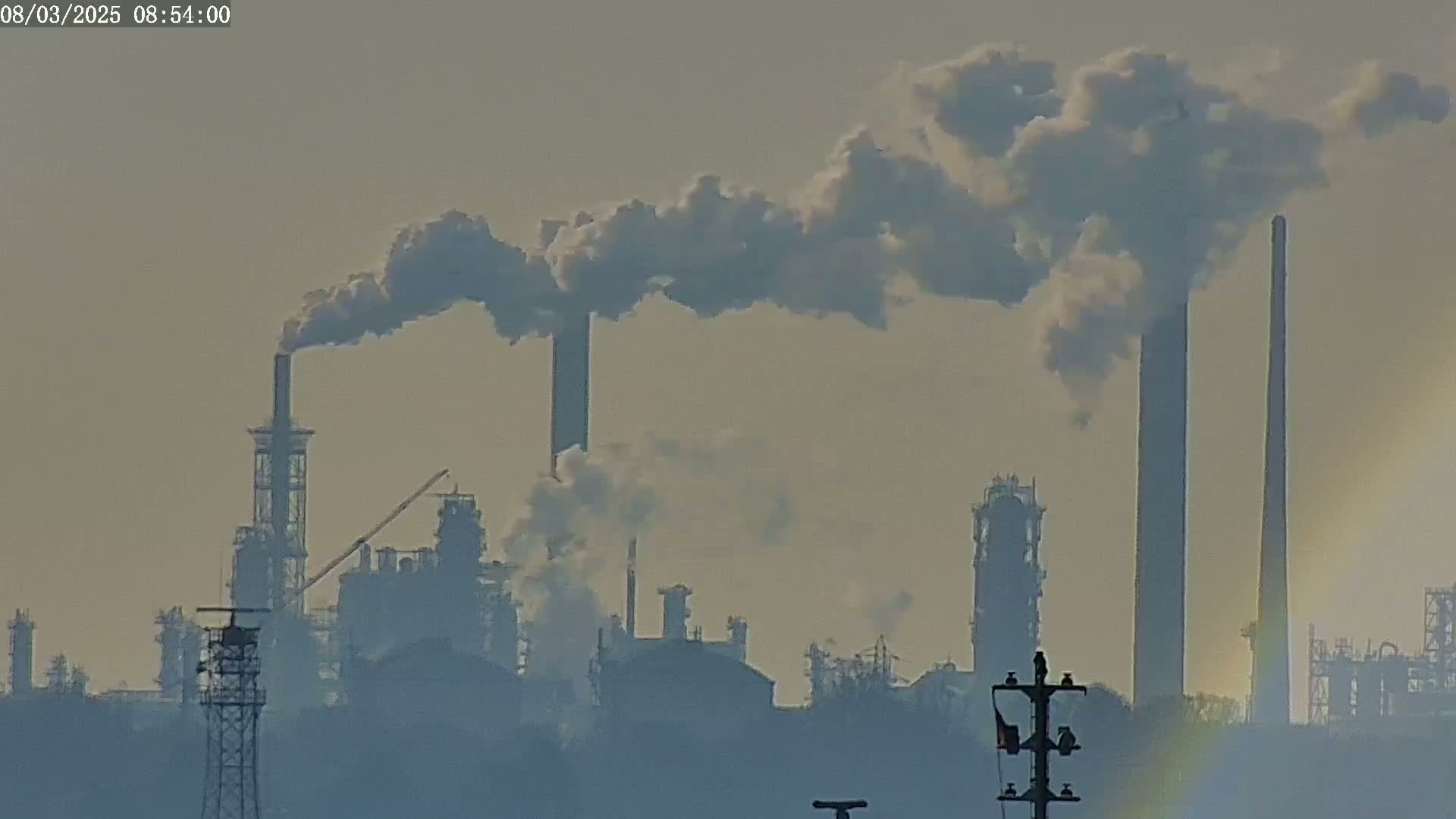 A hazy industrial complex with several tall smokestacks emitting plumes of smoke under a muted sky.