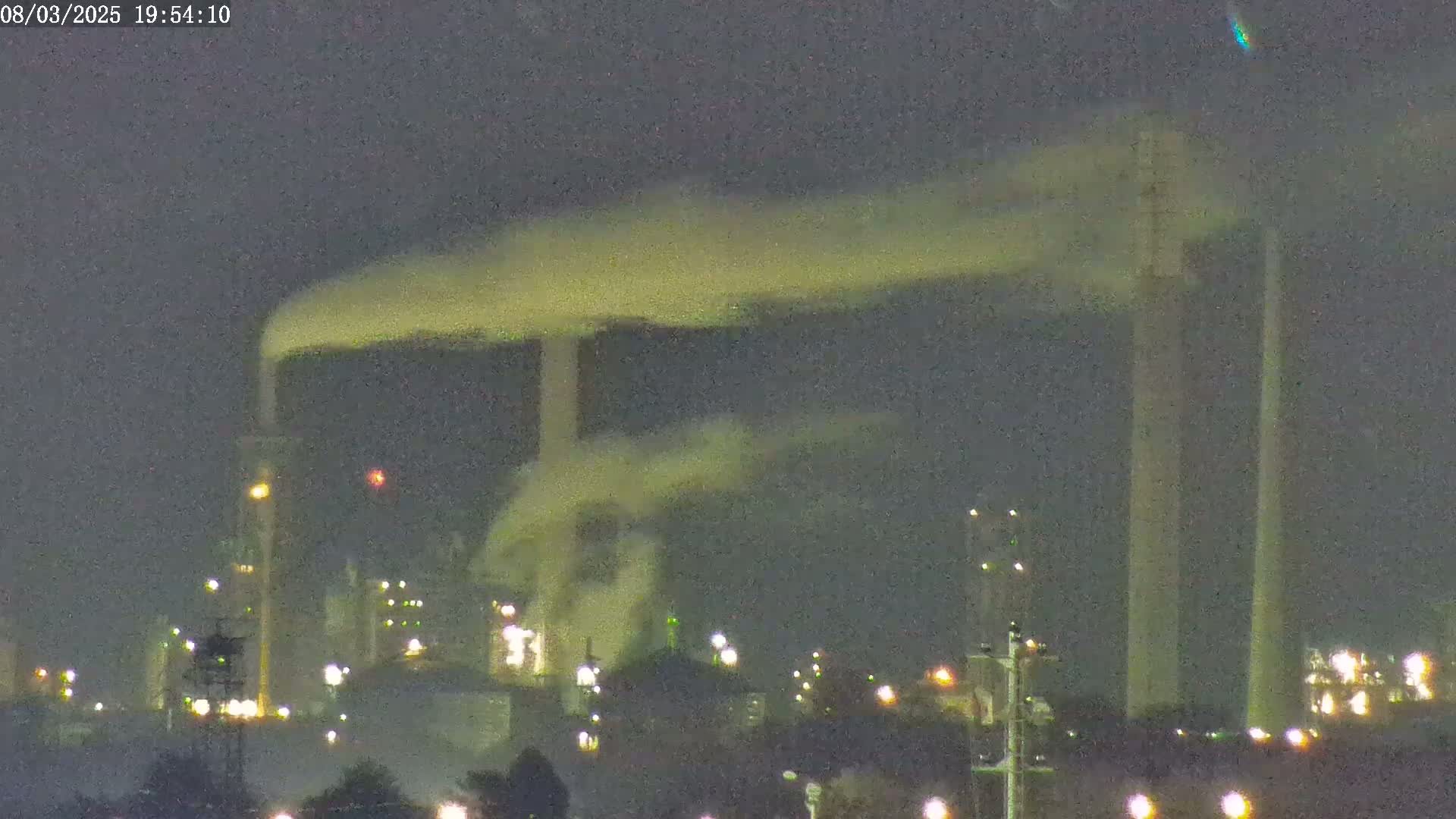 A nighttime view of an industrial plant with tall smokestacks emitting plumes of smoke under a dark sky.