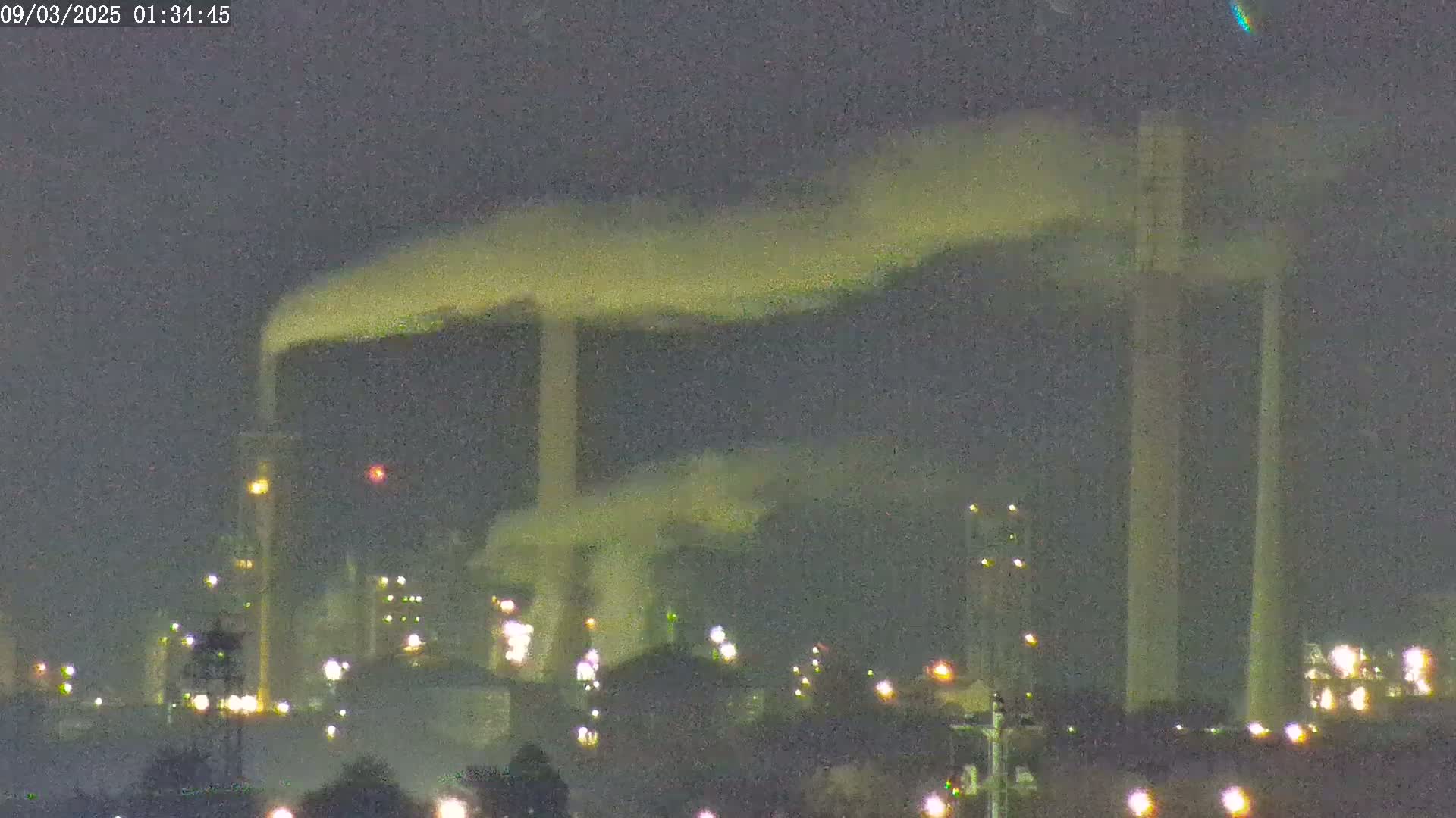 At night, an industrial complex with tall smokestacks emitting plumes of smoke is illuminated by various lights.
