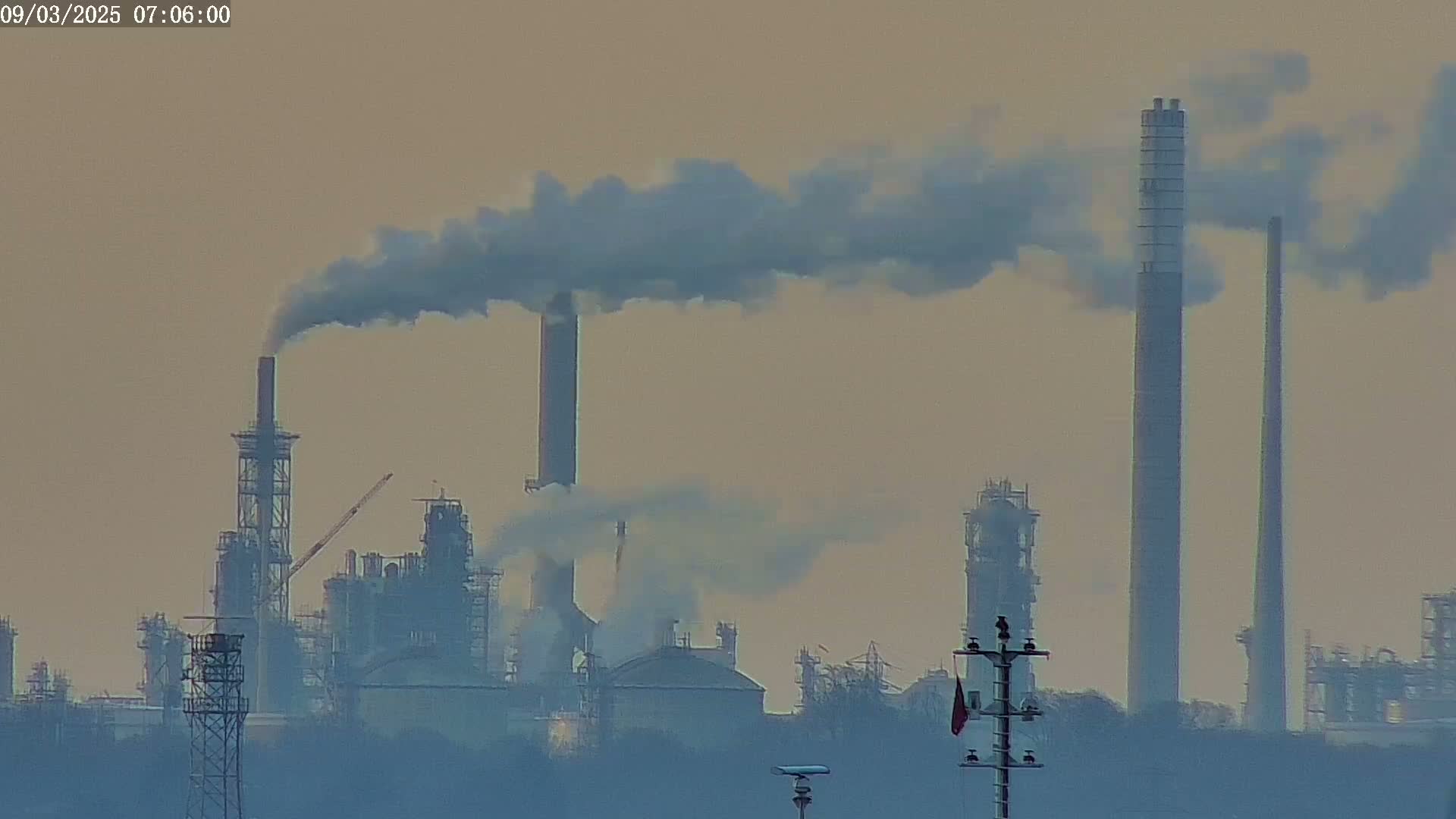 A hazy industrial complex with several tall smokestacks emitting plumes of smoke into a muted, pale sky.