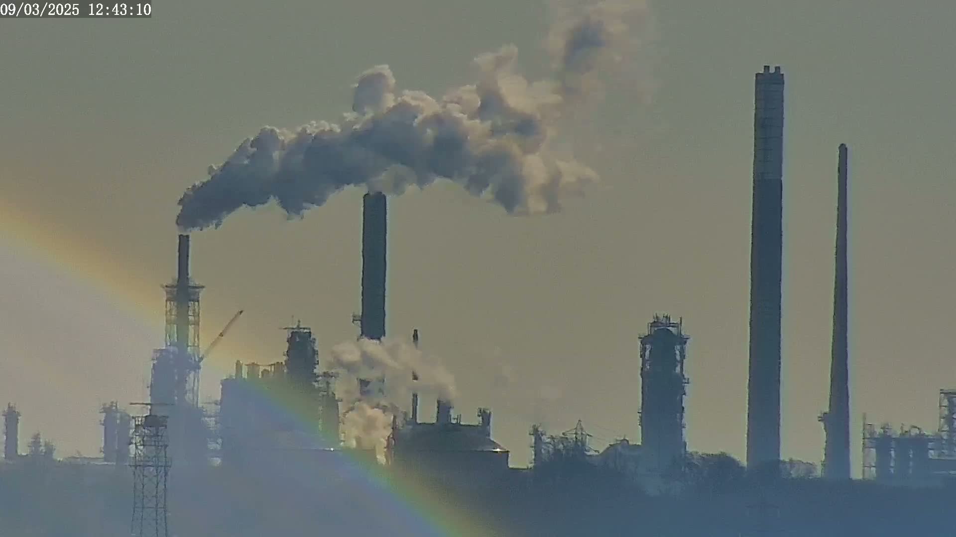 A large industrial complex with several tall smokestacks emitting plumes of smoke under a hazy sky, partially obscured by a faint rainbow.