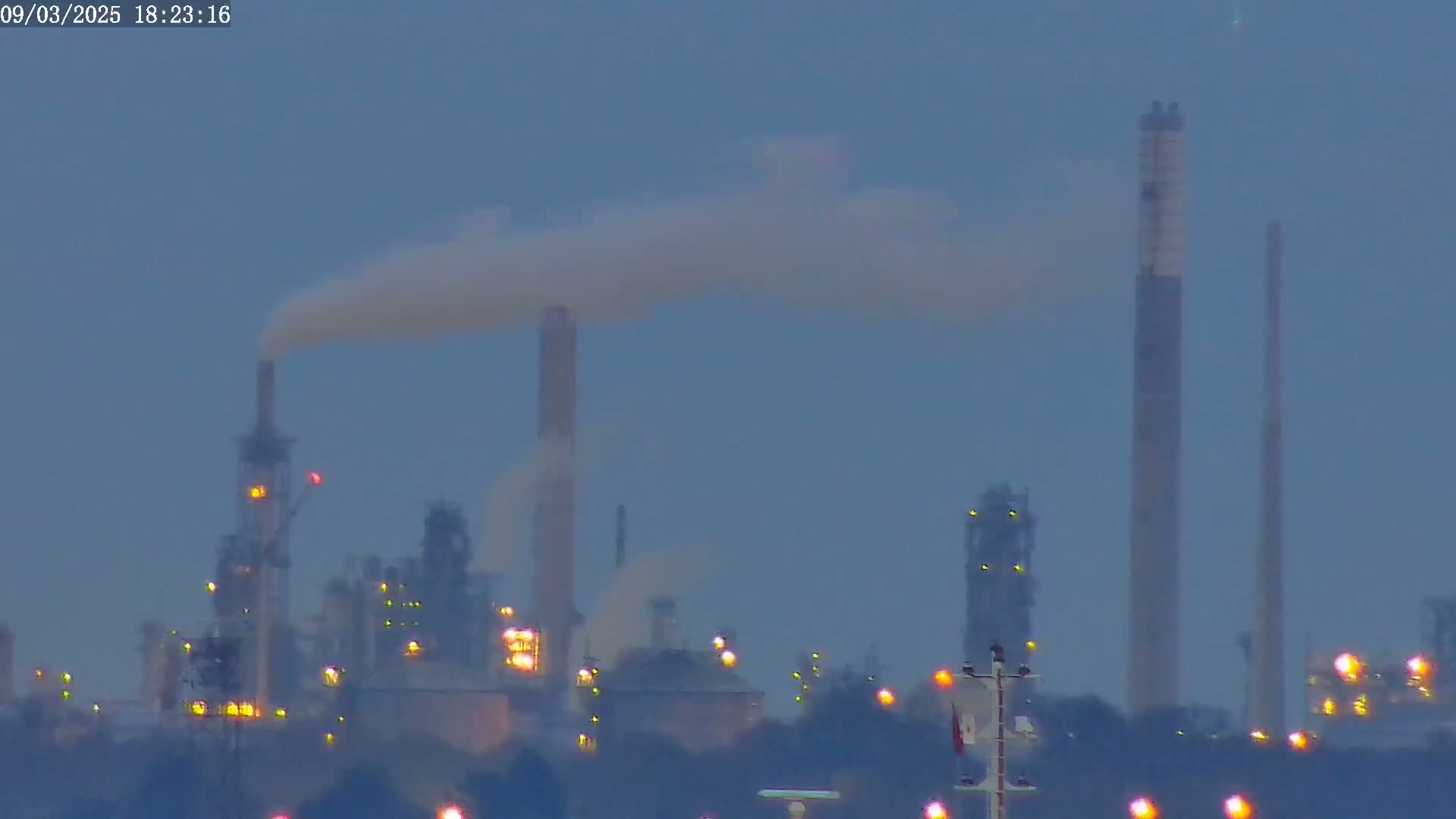 A hazy, dusk-lit industrial complex with several tall smokestacks emitting plumes of smoke.