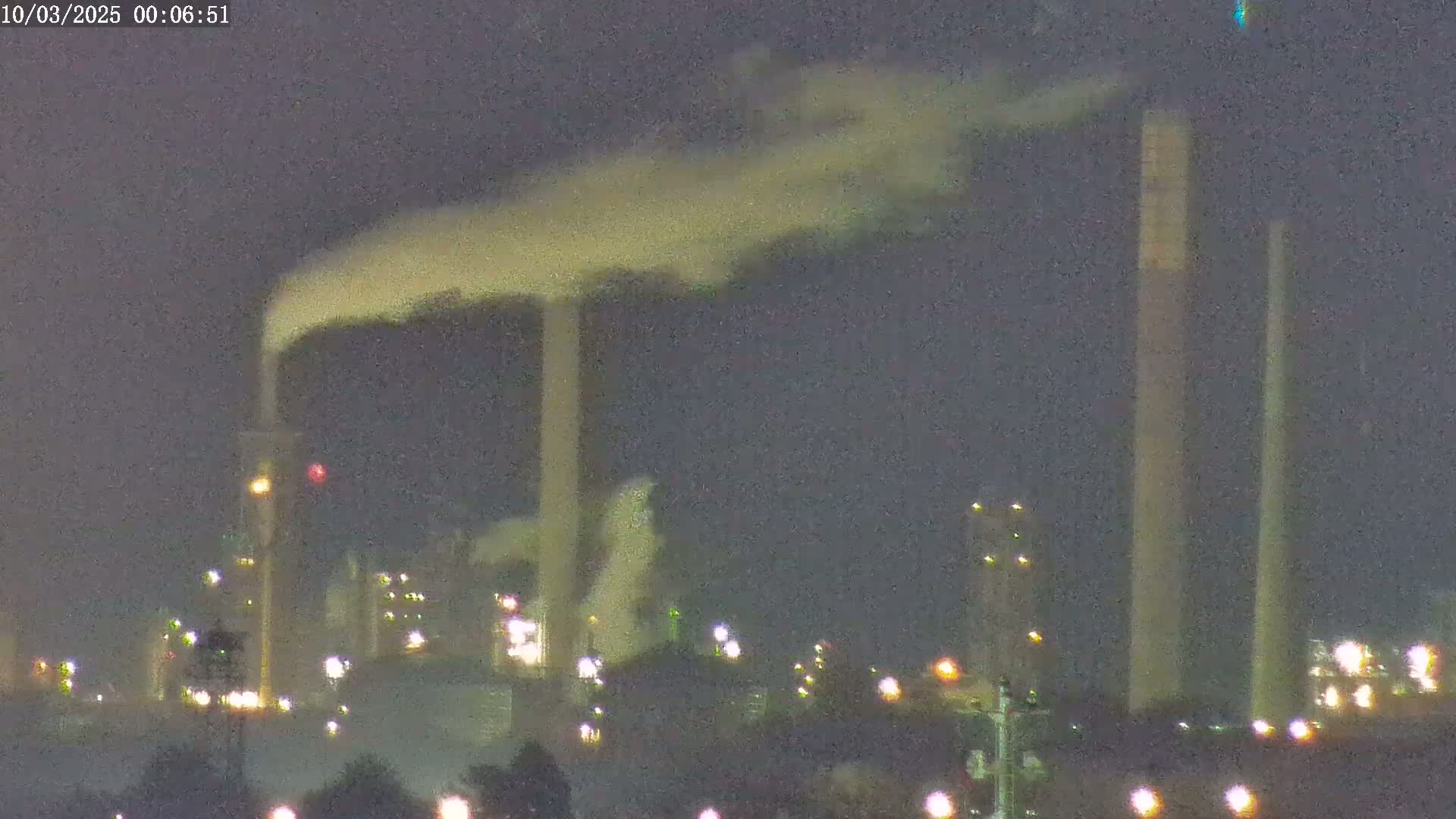 At night, plumes of smoke rise from several tall industrial smokestacks of a factory complex illuminated by scattered lights.