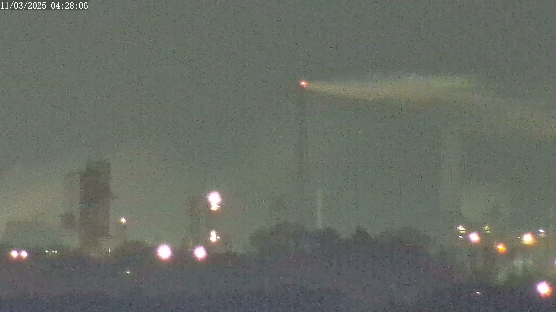 A hazy, nighttime view shows an industrial complex with tall structures and several lights, one of which is emitting a bright beam of light into the atmosphere.