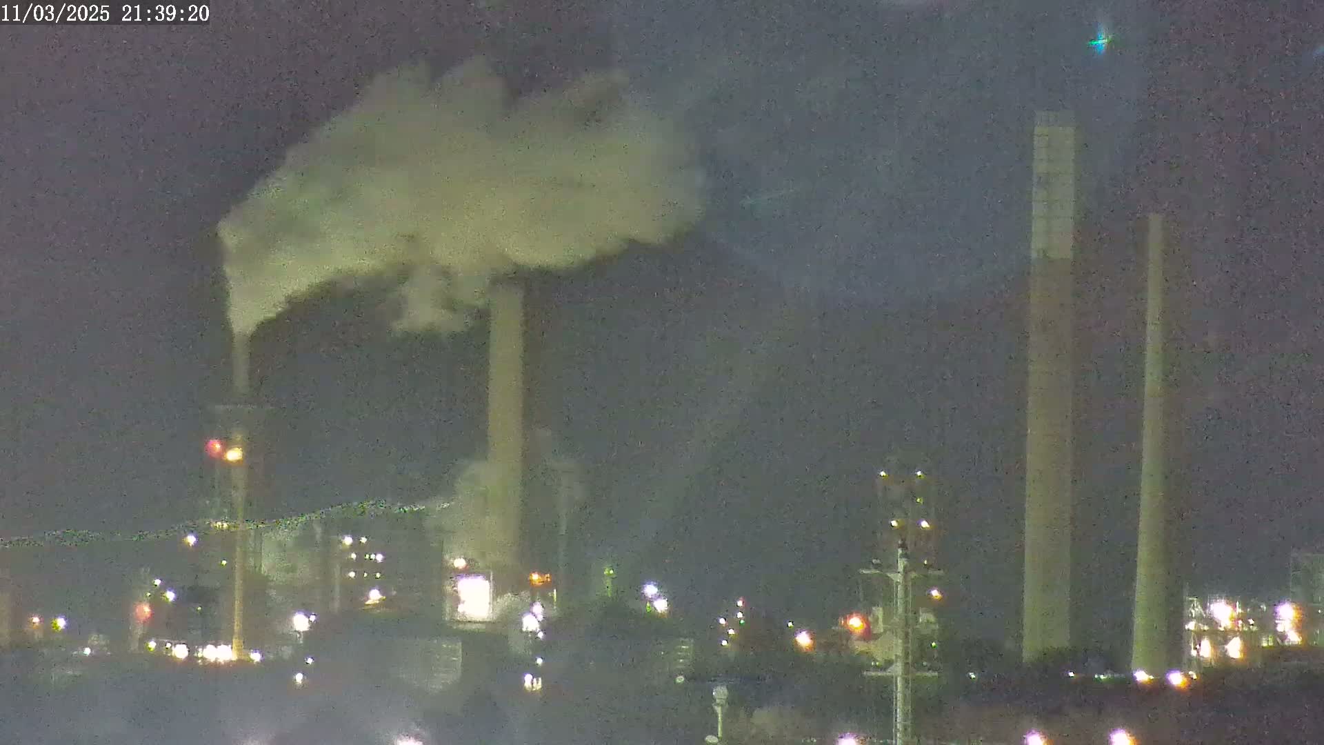 At night, an industrial plant with tall smokestacks emits a large plume of smoke under a dark sky.