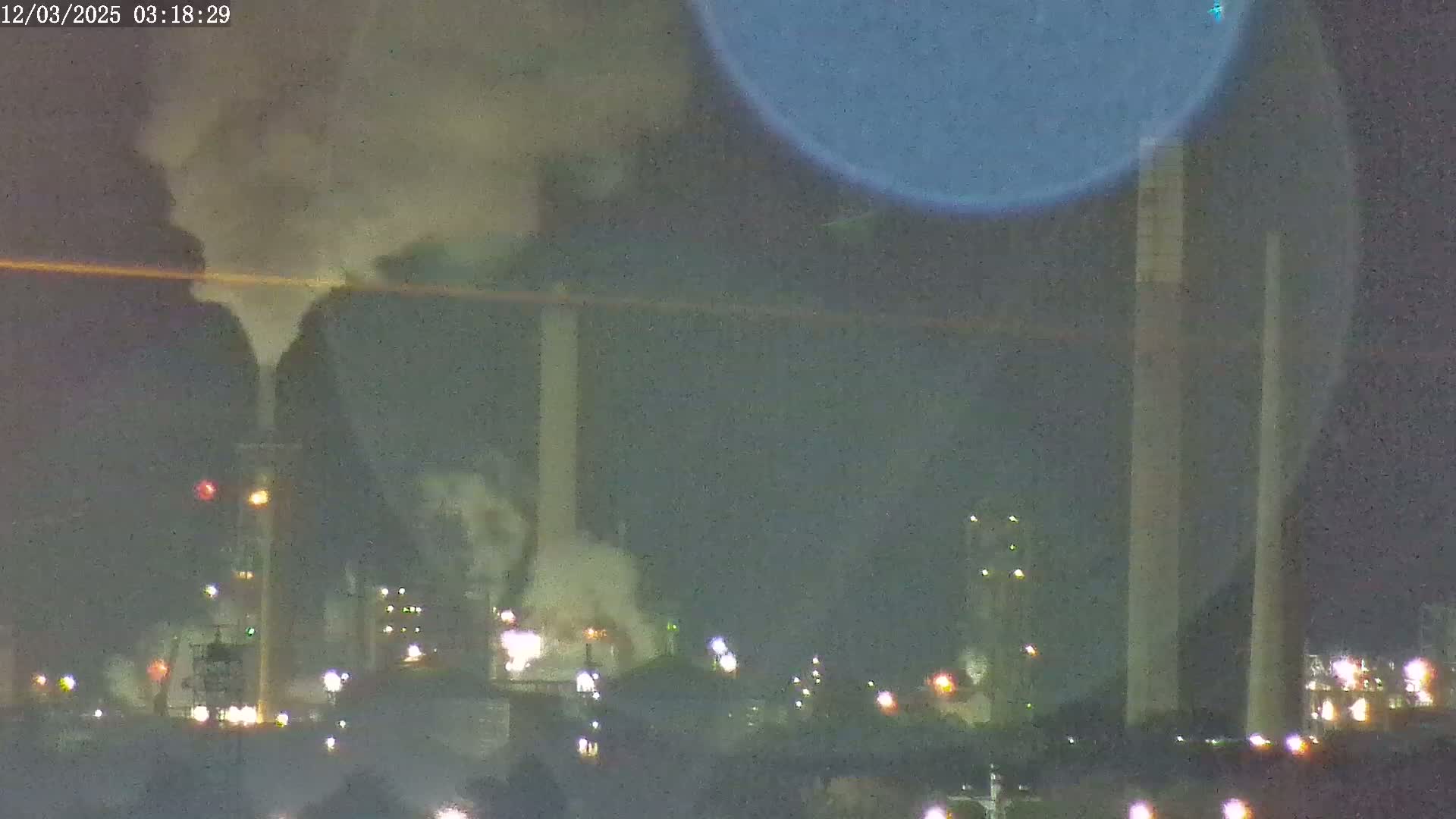 A nighttime view of an industrial complex emitting smoke into a dark, hazy sky.