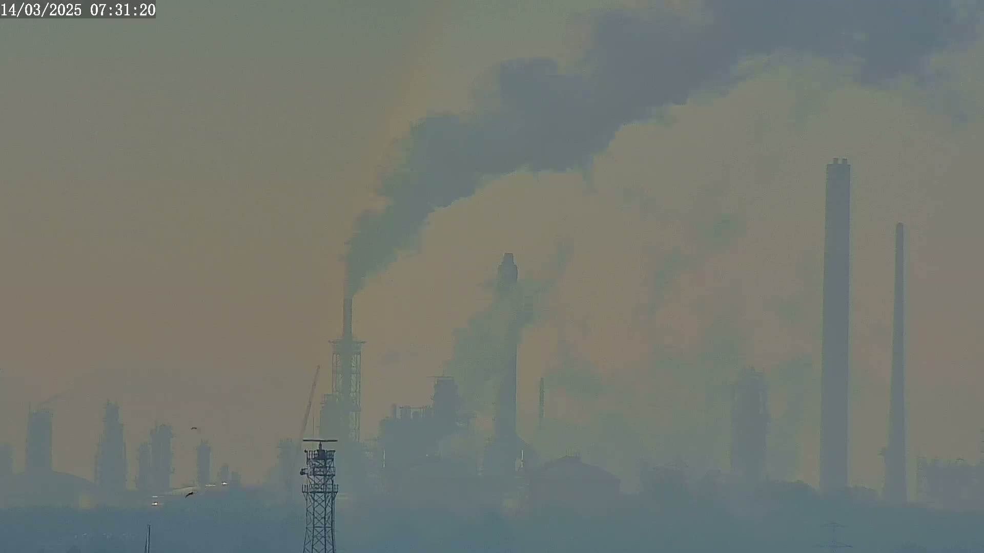 A hazy, smoggy view shows an industrial plant with several tall smokestacks emitting dark plumes of smoke into the air.