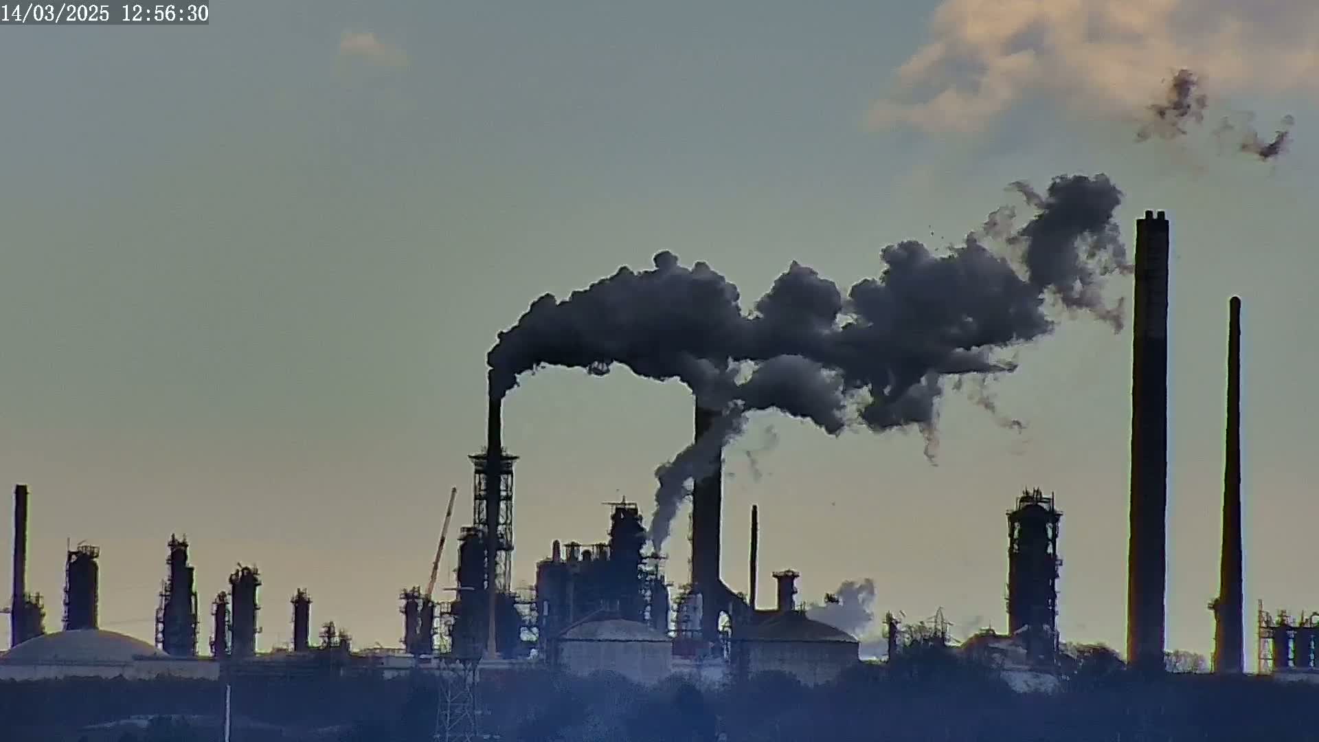 A large industrial plant with several tall smokestacks emitting dark plumes of smoke into a partly cloudy sky.