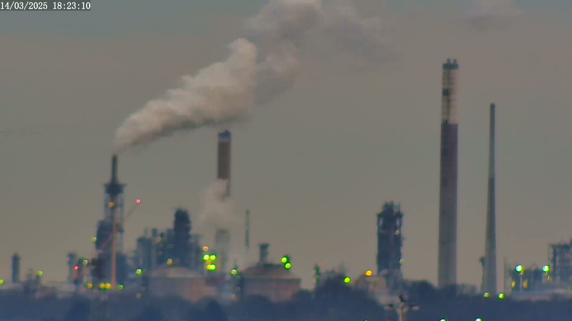 A distant industrial complex with several tall smokestacks, one emitting a plume of white smoke, under a hazy sky.