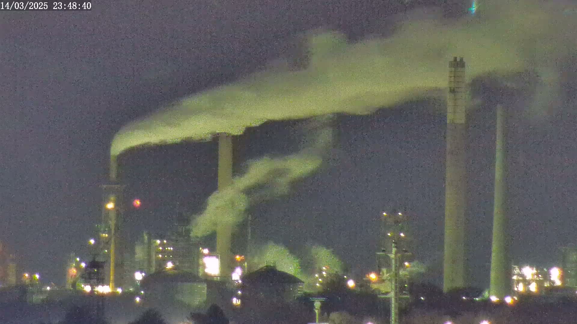 At night, plumes of smoke rise from several tall industrial smokestacks above a brightly lit factory complex.