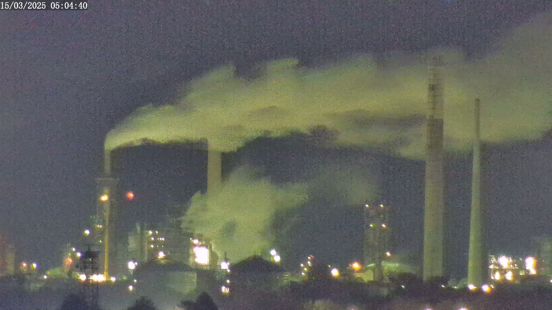 At night, an industrial complex emits plumes of yellowish-green smoke into a dark sky.