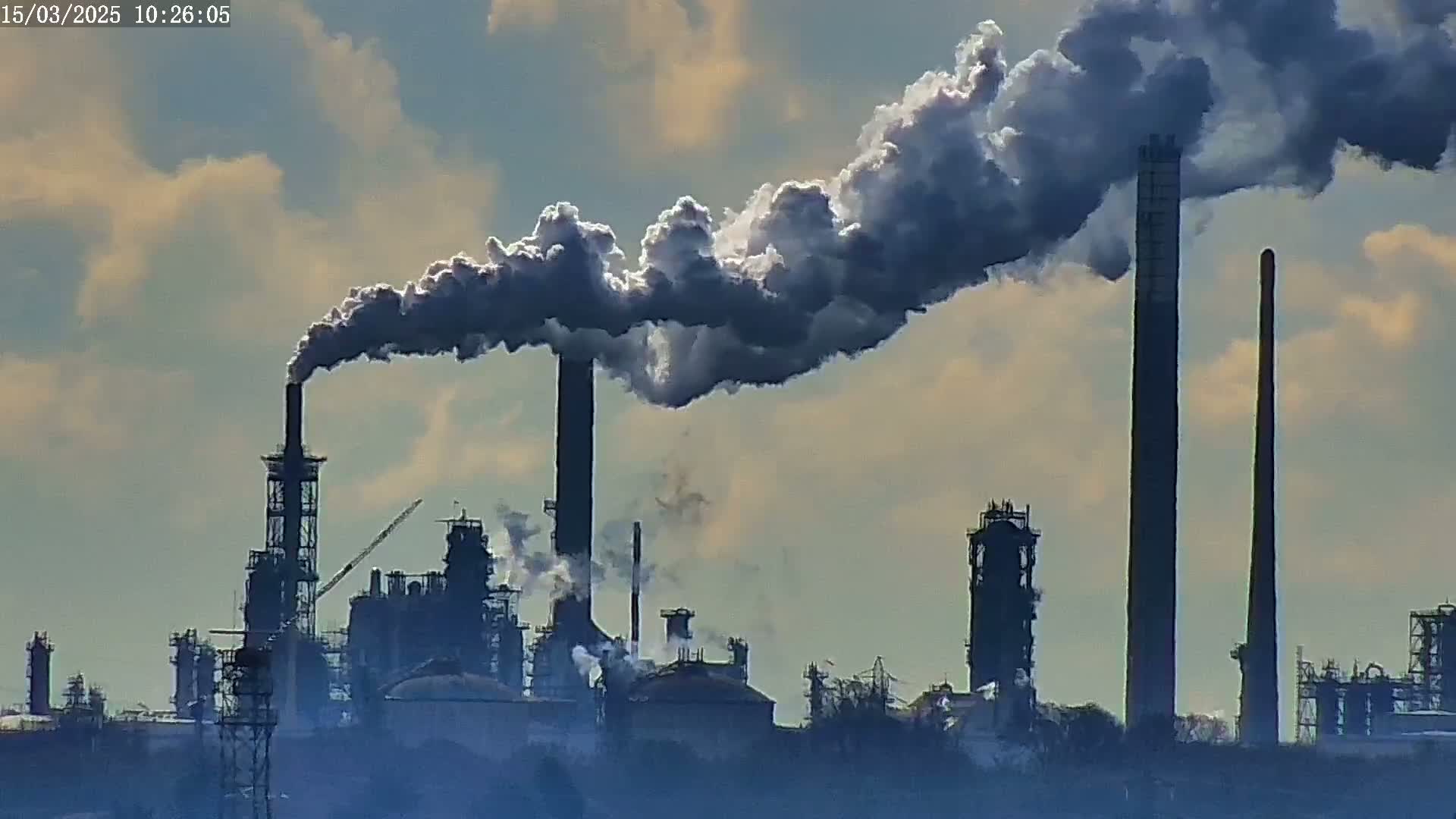 A large industrial plant releases plumes of dark smoke into a partly cloudy sky.